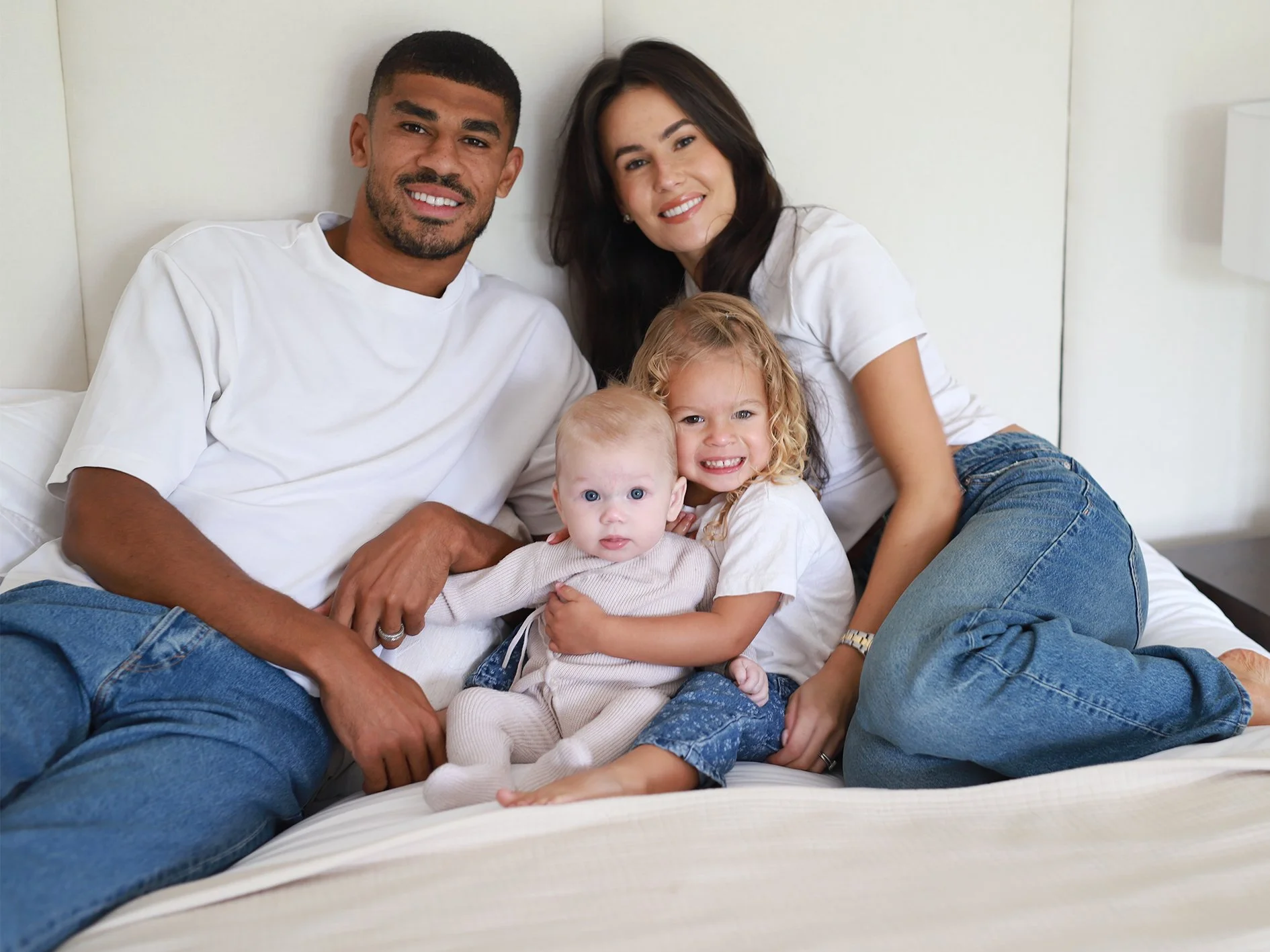 A family of four sitting on a bed, smiling at the camera. The group includes an adult man and woman, and two young children, one with curly blonde hair and the other with short blonde hair.