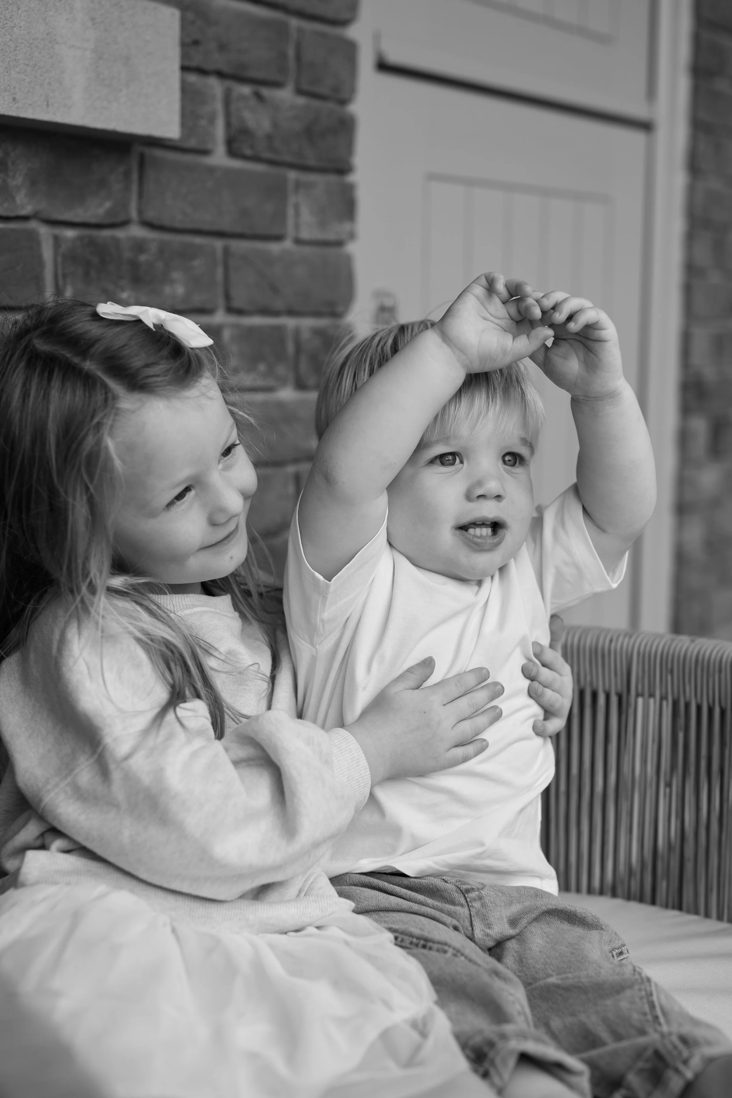 A young girl and a young boy sitting together, with the girl hugging the boy. The girl is smiling and looking at the boy, who is reaching up with his arms raised. The scene is in black and white.