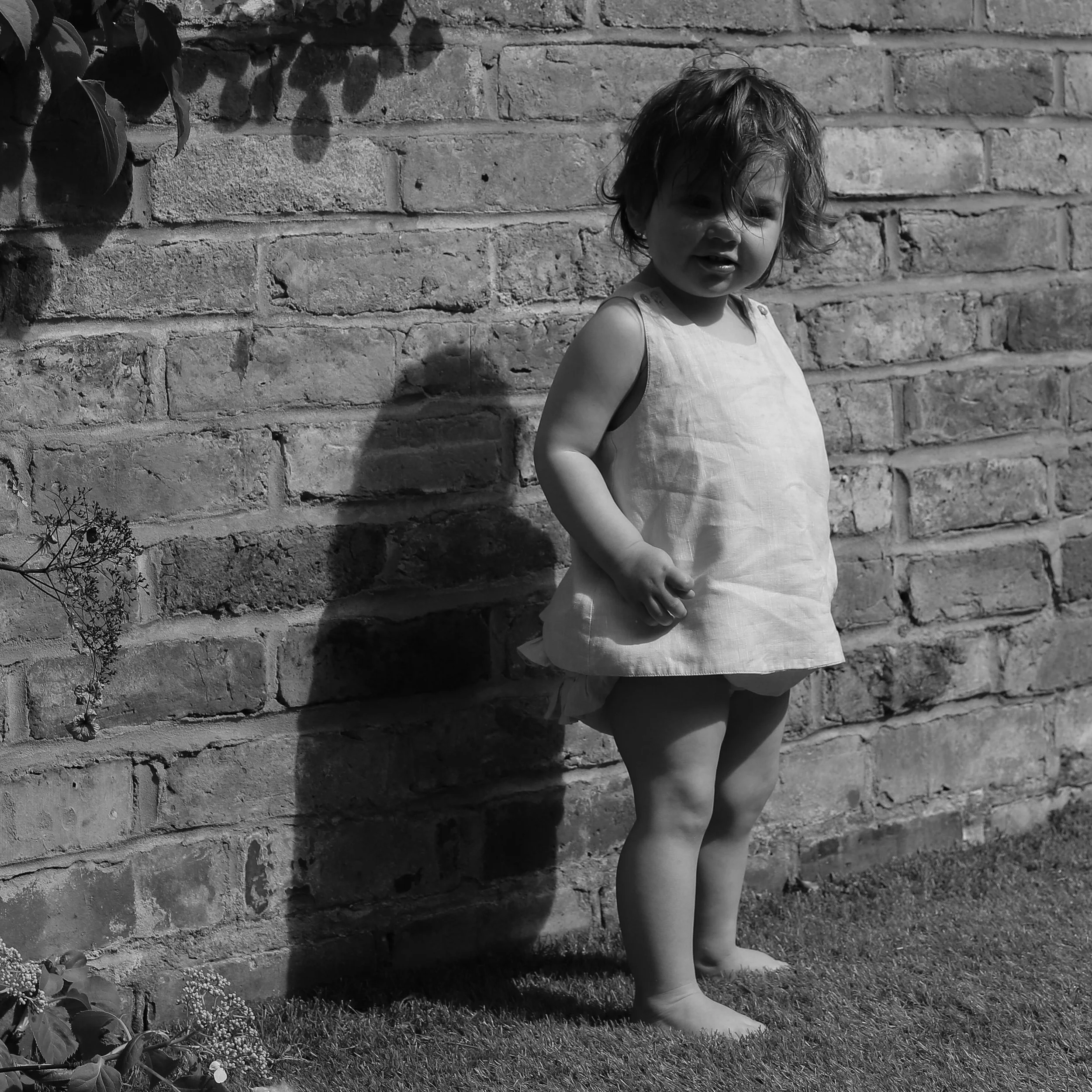 A young girl with messy hair standing barefoot on grass in front of a brick wall, casting a shadow.