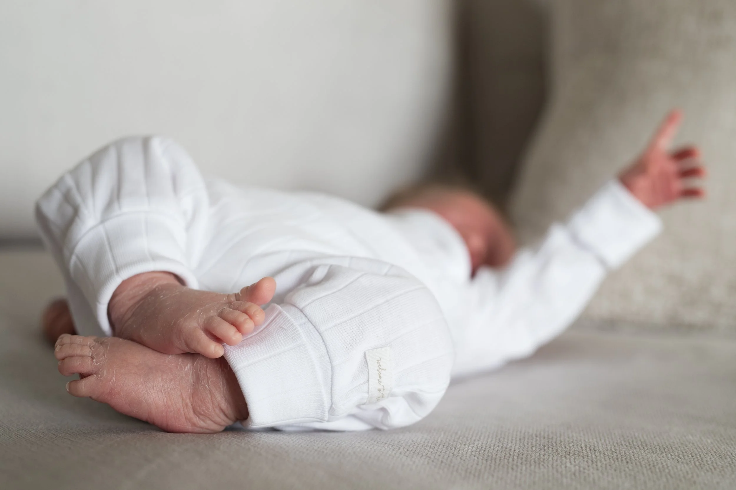 A newborn baby lying face down on a bed wearing a white outfit, with a small hand resting on a larger adult hand.