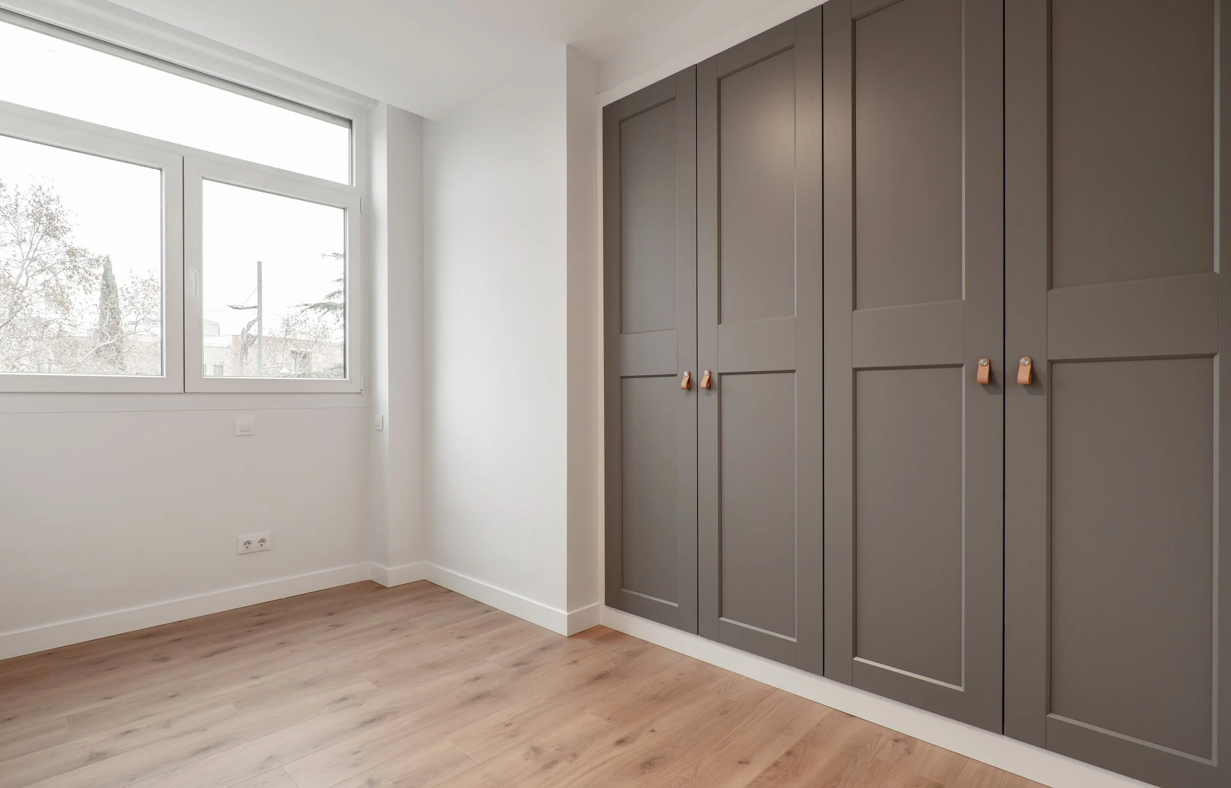 Empty room with large window, white walls, brown wooden floor, and grey closet with brown handles.