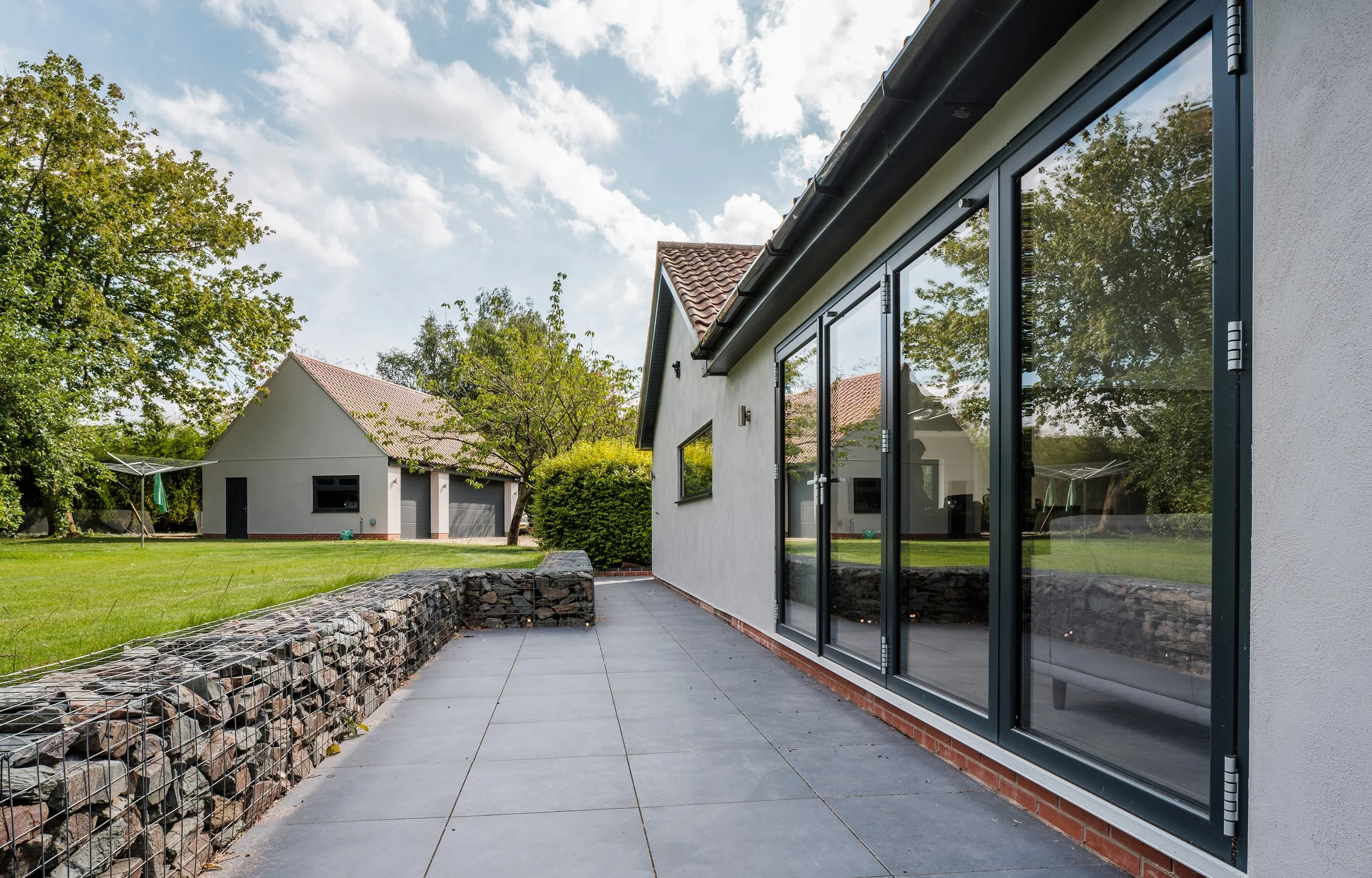 A backyard patio with large sliding glass doors on a white house, a stone wall, green lawn, trees, and a detached garage in the background on a partly cloudy day.