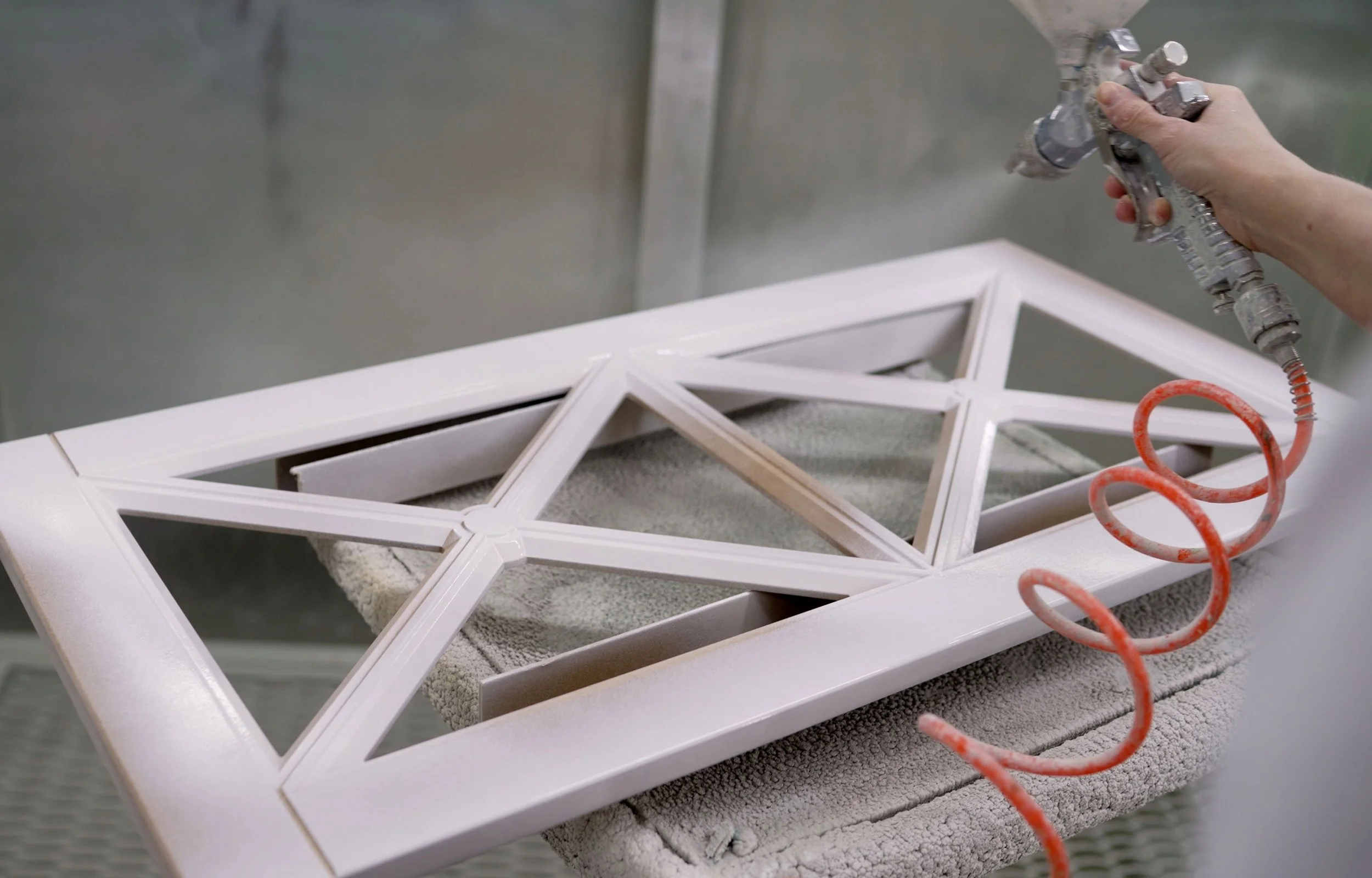 Person spray painting a white wooden picture frame with geometric design in a workshop.
