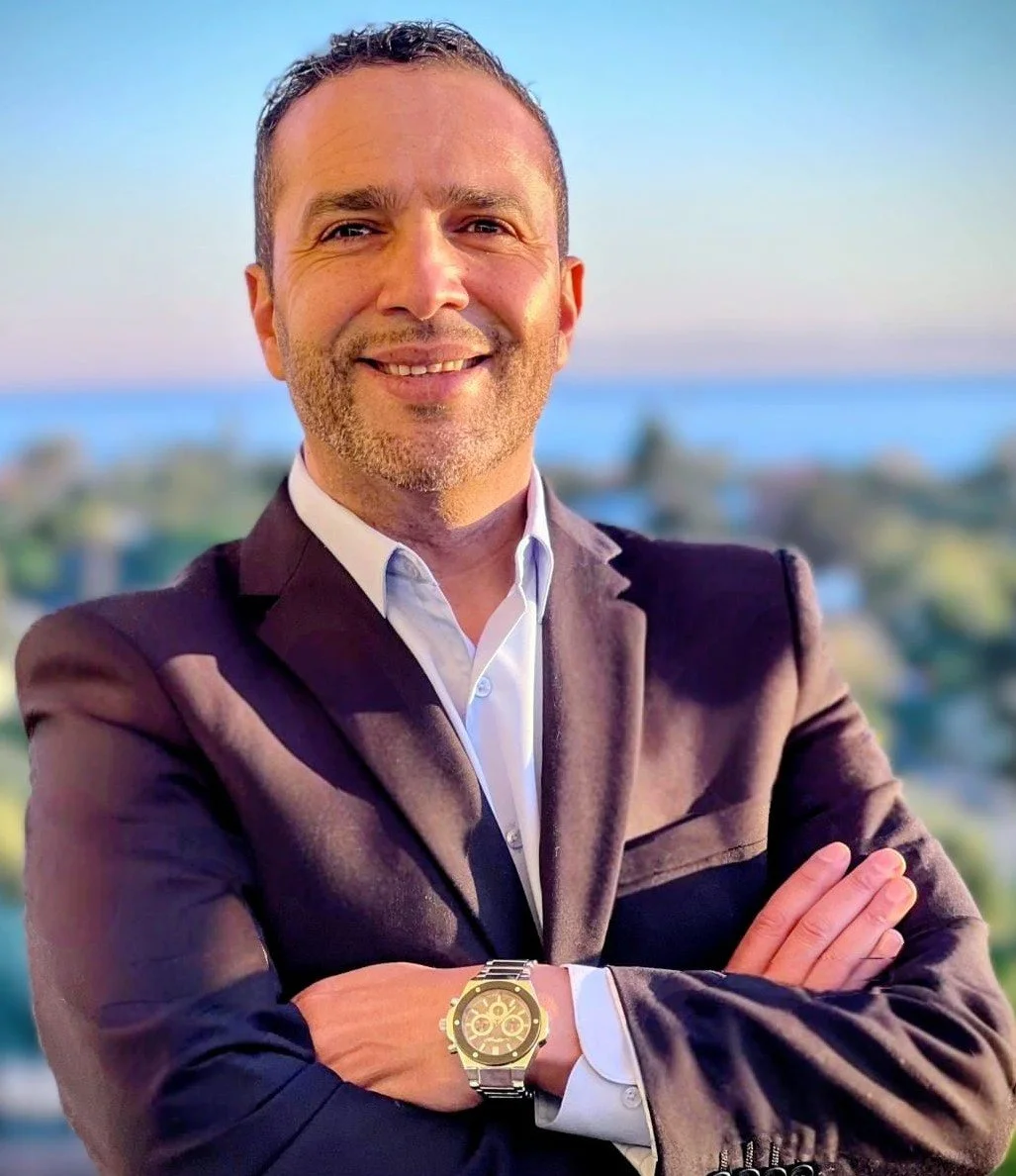A smiling man in a suit and white shirt with arms crossed, standing outdoors with a blurry scenic background of water and landscape.
