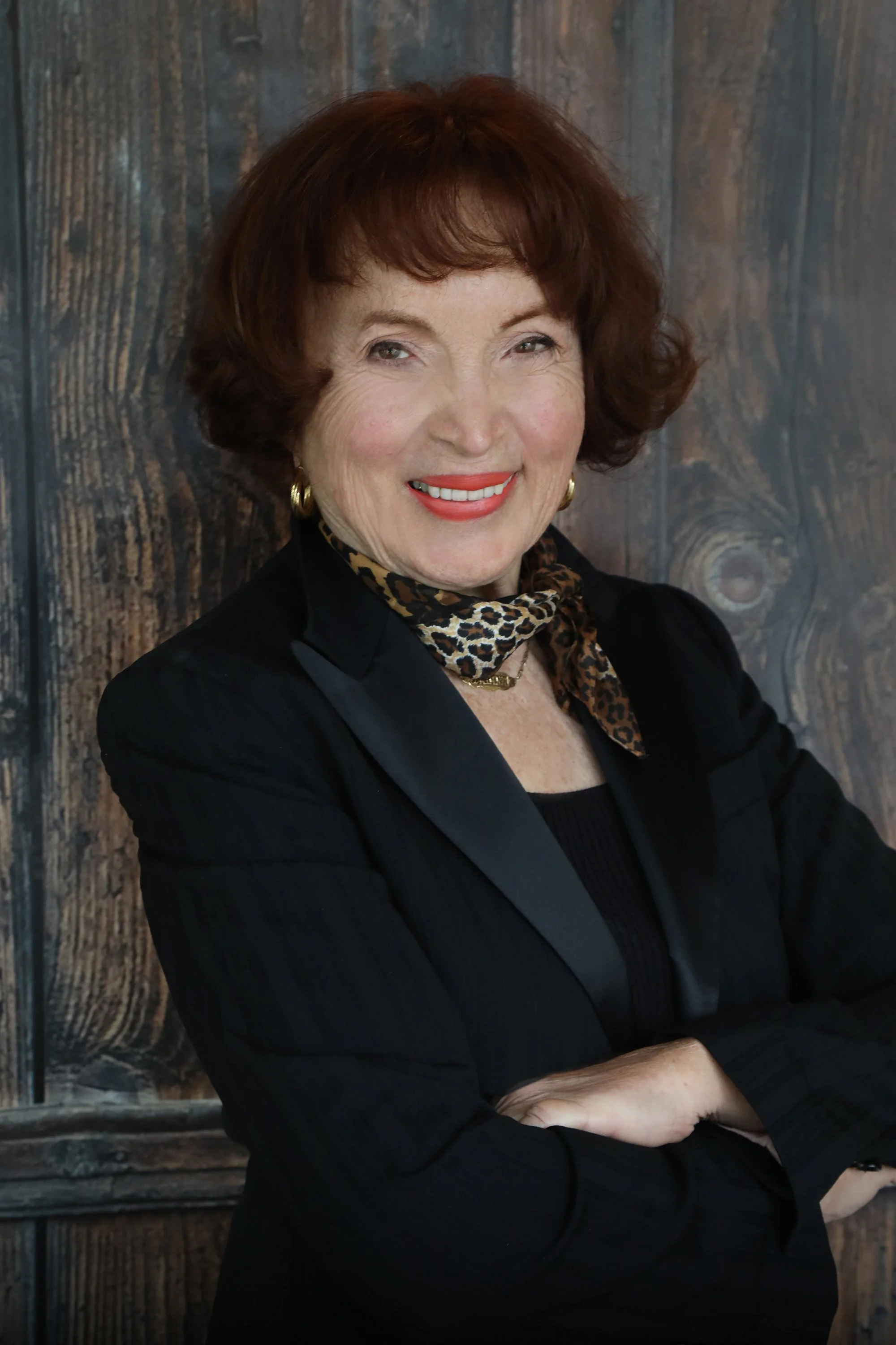Portrait of an elderly woman with short brown hair, wearing a black blazer, a leopard print scarf, and gold earrings, smiling and standing with arms crossed.