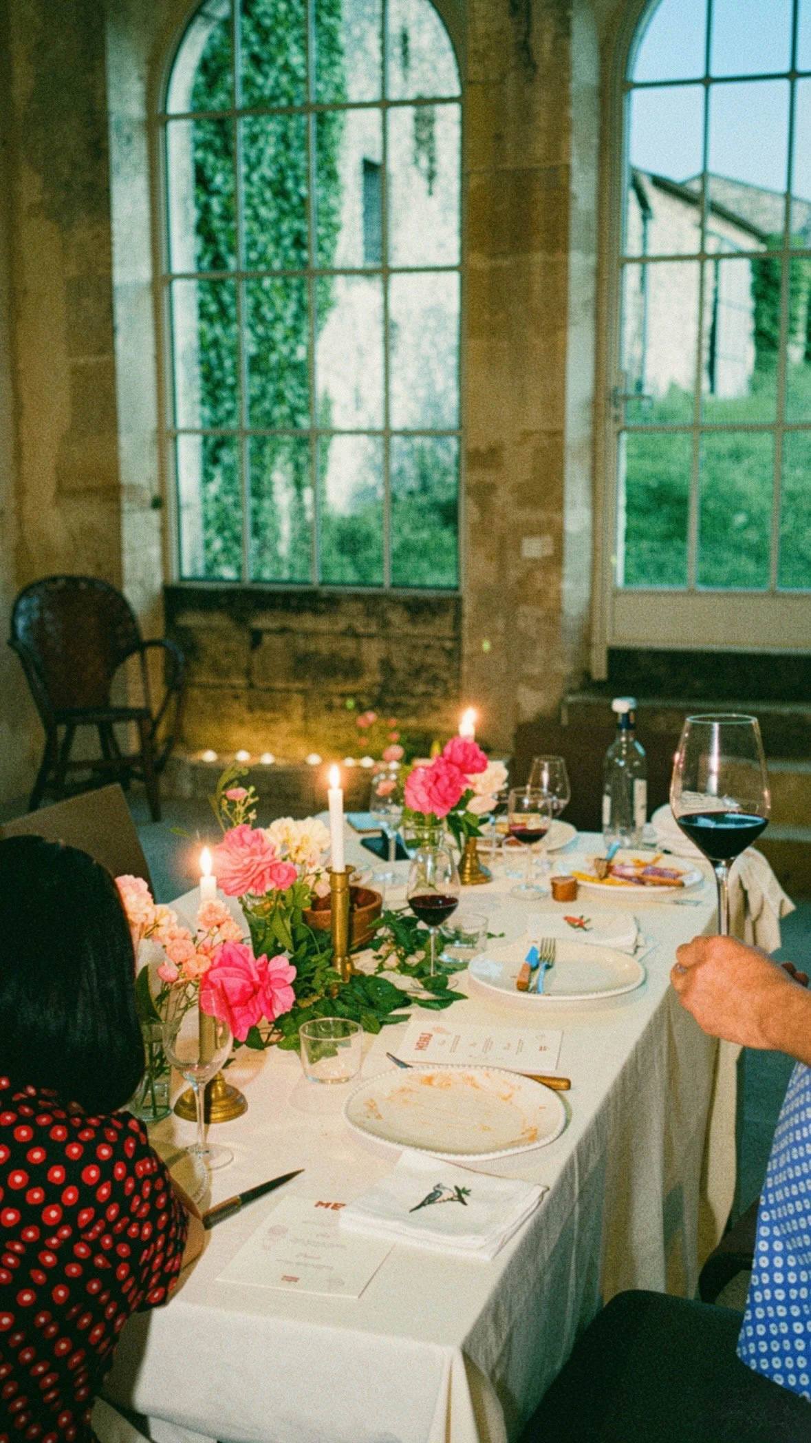 Table de fête avec des fleurs roses, des bougies, des verres de vin rouge, de l'eau, assiettes, et des invités lors d'une célébration dans une salle lumineuse avec de grandes fenêtres.