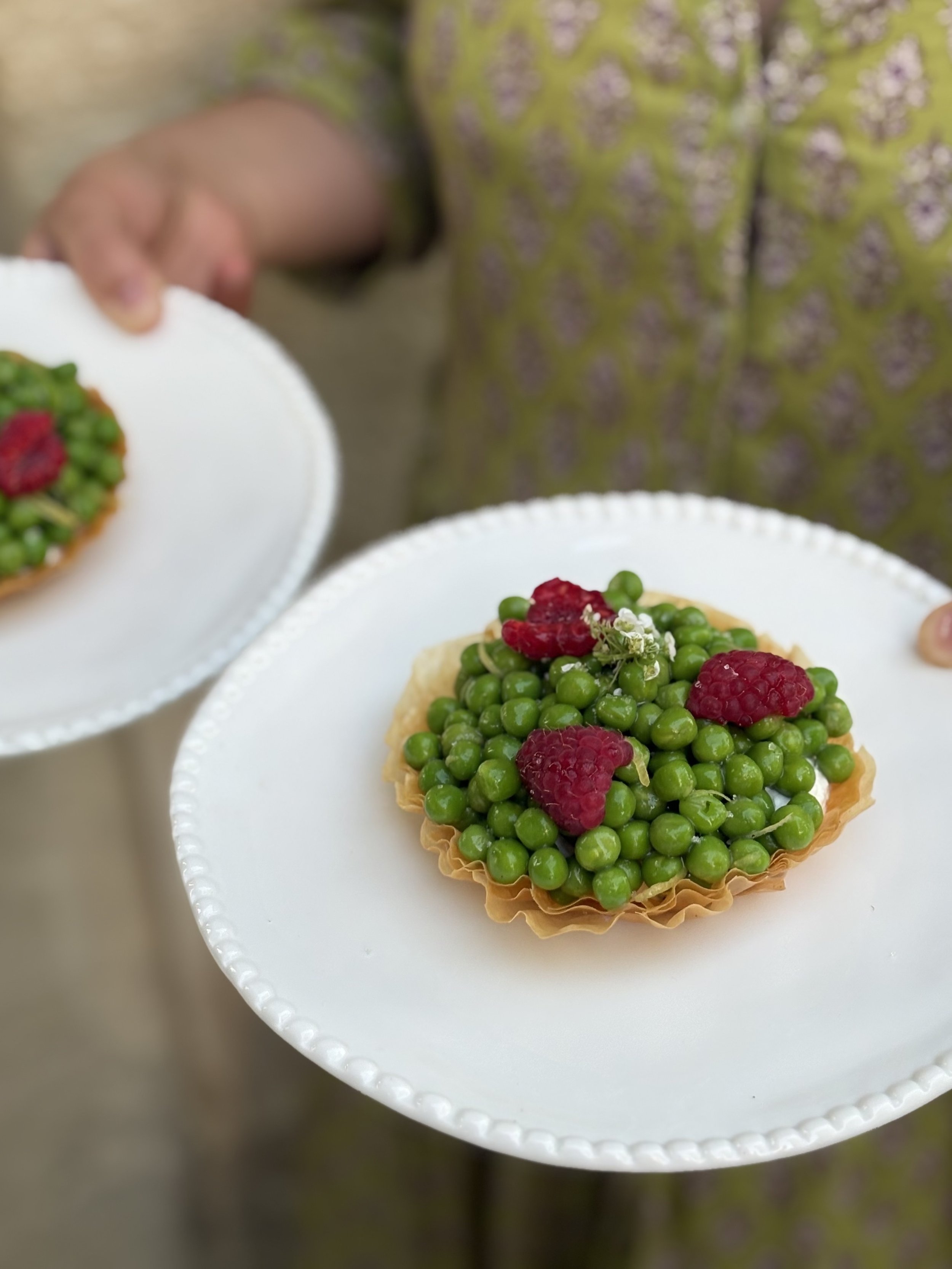 Une tartelette aux pois verts et framboises sur une assiette blanche, avec une personne en arrière-plan portant un vêtement vert avec motifs floraux.