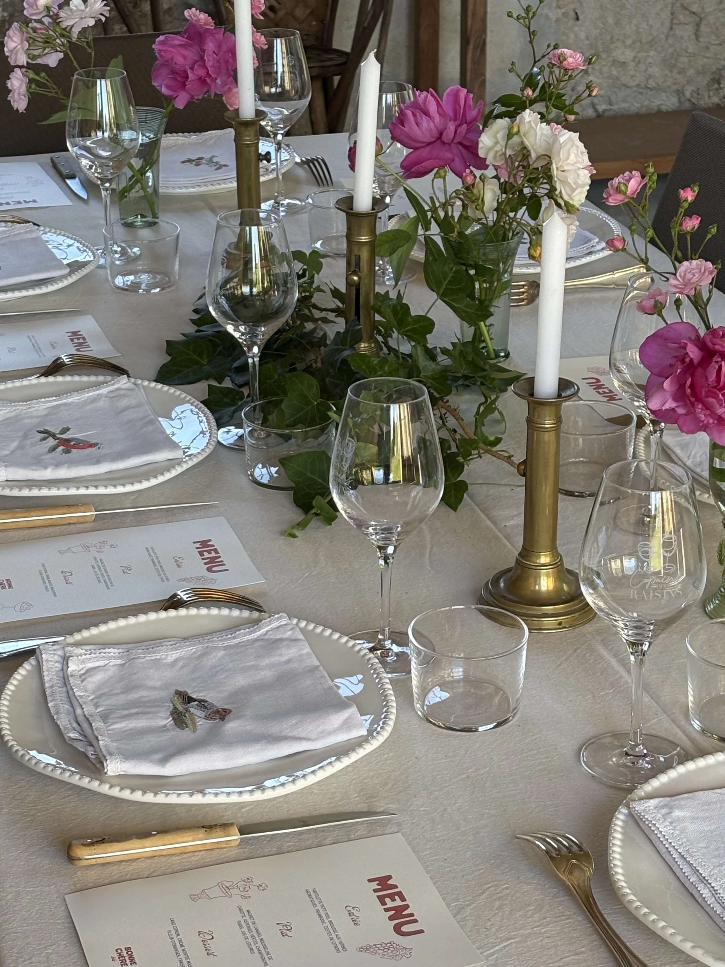 Une table élégante dressée pour un repas, décorée avec des fleurs roses et blanches dans un vase, des bougies blanches dans des chandeliers, des assiettes avec des serviettes pliées, des verres à vin et des couverts en argent, et un menu écrit.
