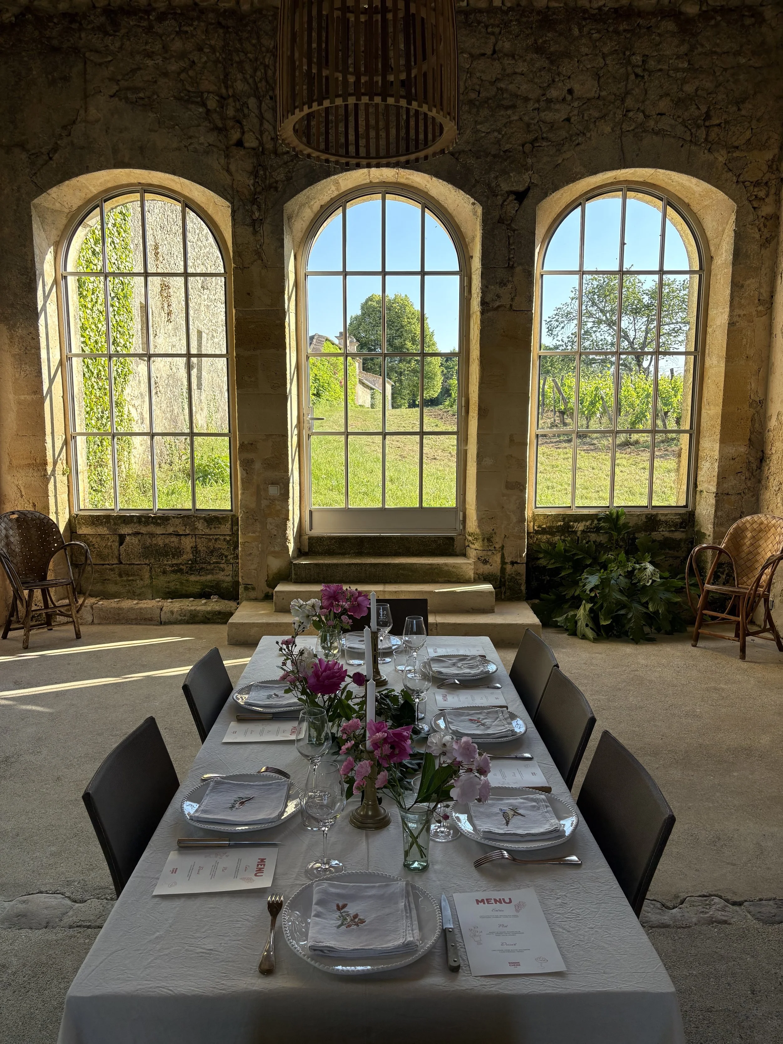 Table dressée avec des assiettes, des verres, des couverts et un centre de table avec des fleurs, dans une salle avec trois grandes fenêtres donnant sur un jardin ensoleillé.