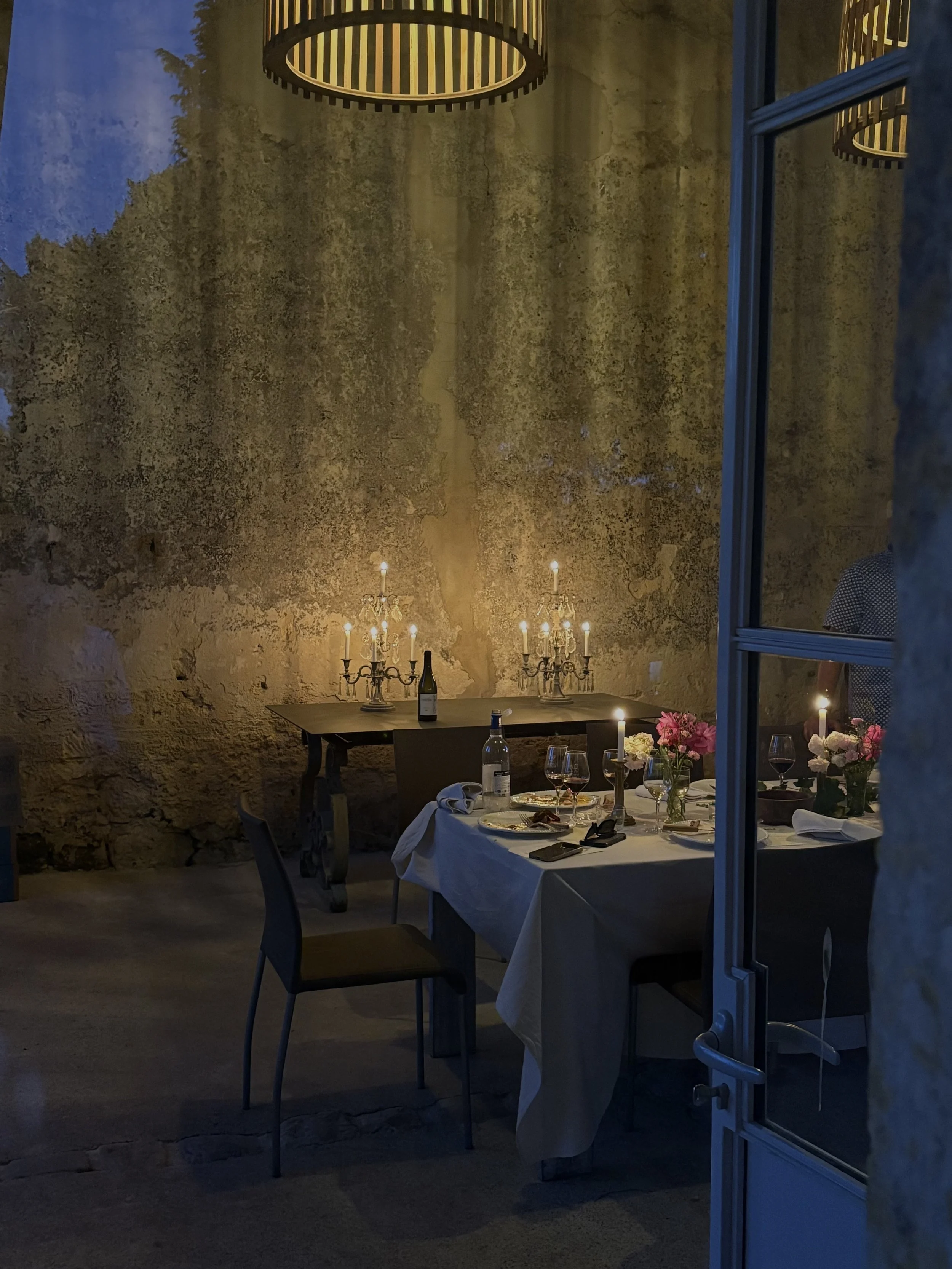 Une salle de dîner élégante avec un mur en pierre, une table est dressée avec une nappe blanche, des chandeliers avec des bougies allumées, des fleurs, des verres à vin et une bouteille de vin.