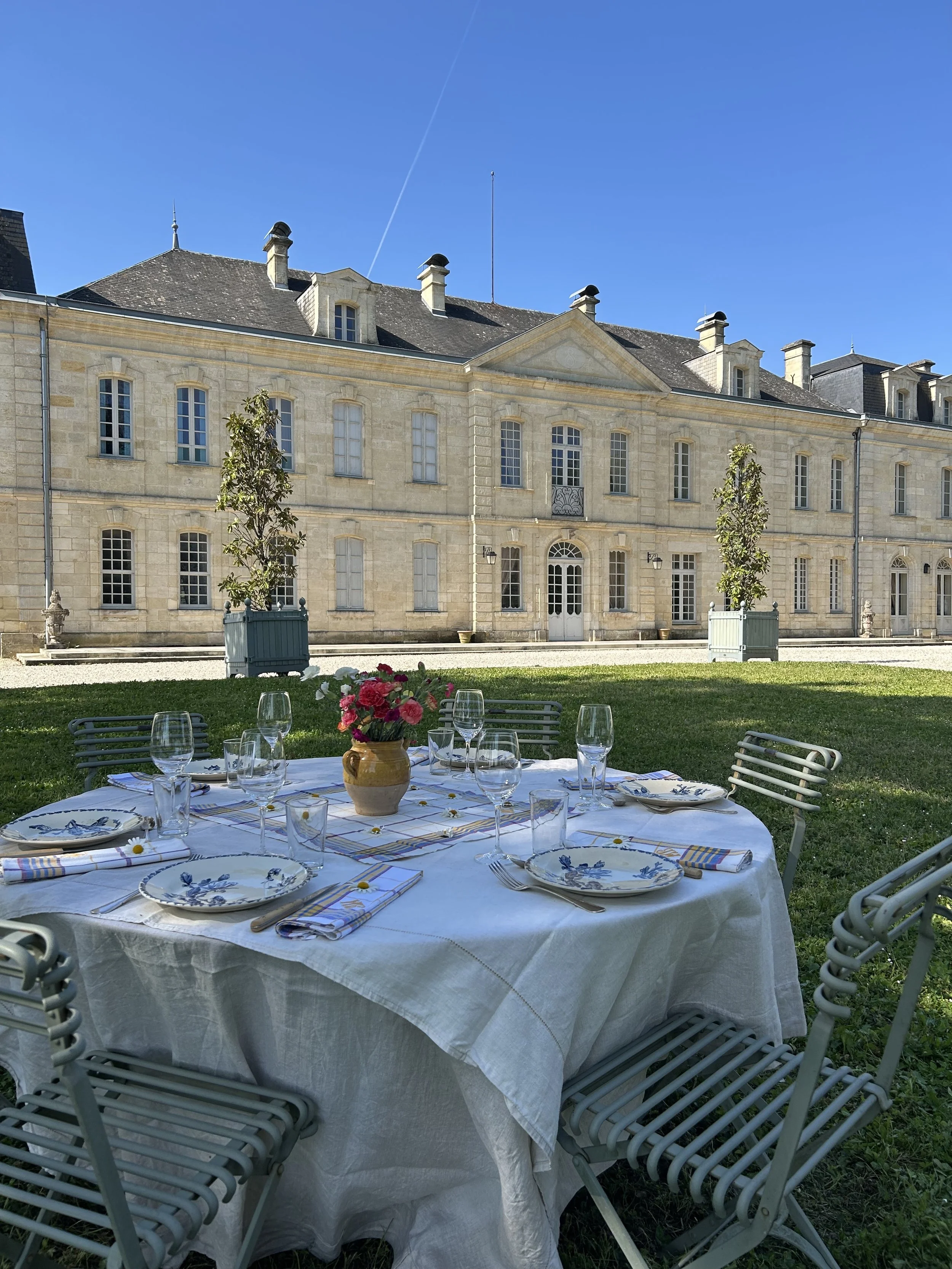 Table dressée pour un repas en extérieur avec des verres, des assiettes et un vase de fleurs, face à un grand bâtiment en pierre dans un jardin ensoleillé sous un ciel bleu.