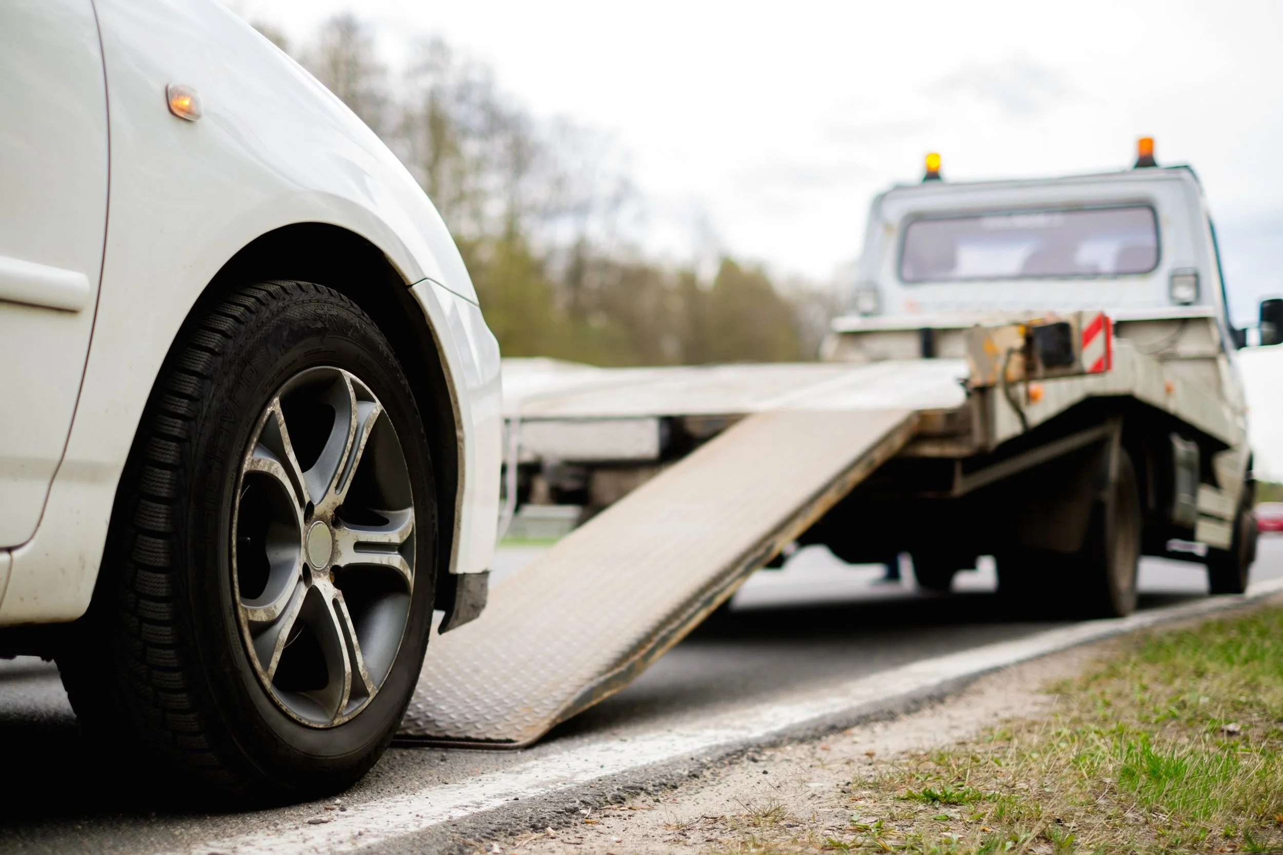 A white tow truck with an empty flatbed is towing a white car with a matching alloy wheel from a roadside.