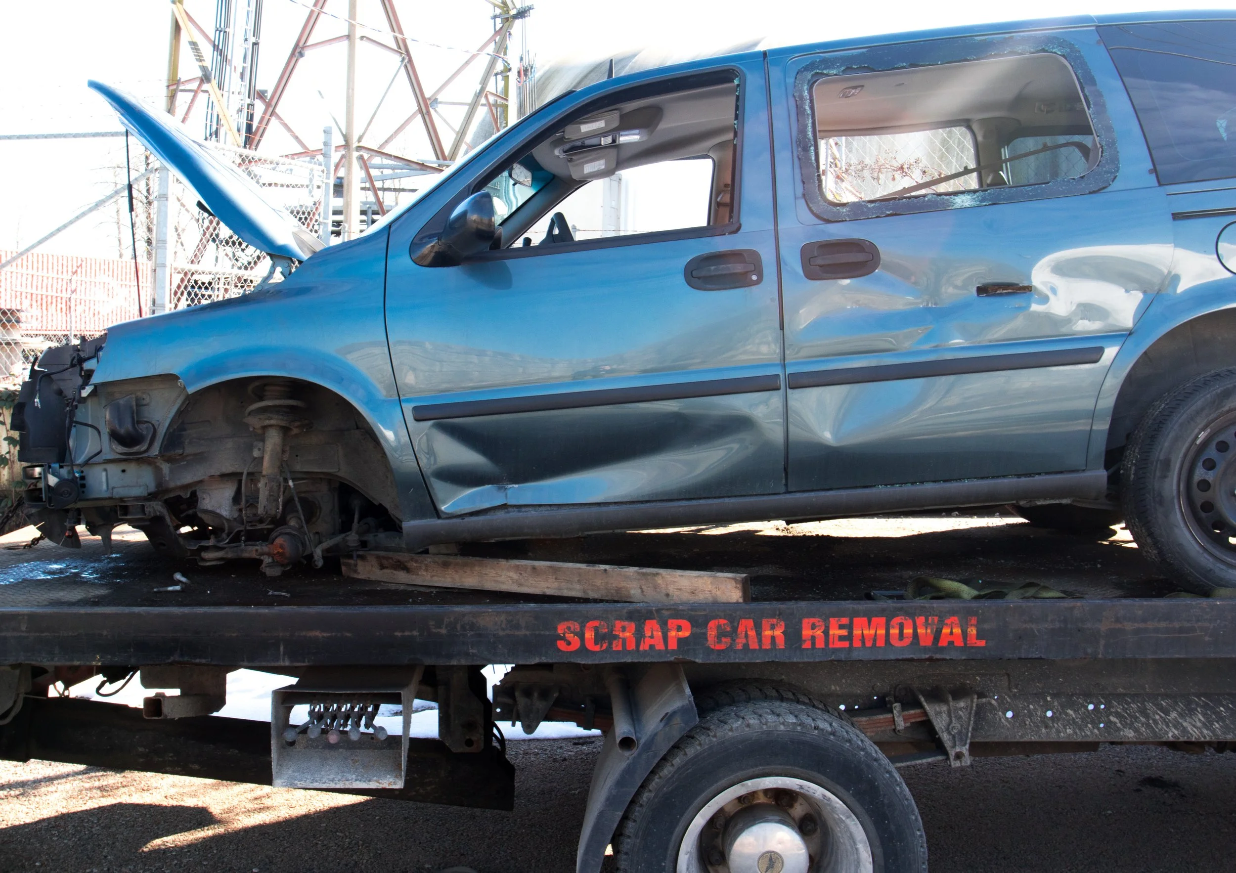 A blue car with front damage is on a flatbed tow truck with 'SCRAP CAR REMOVAL' written on the side.