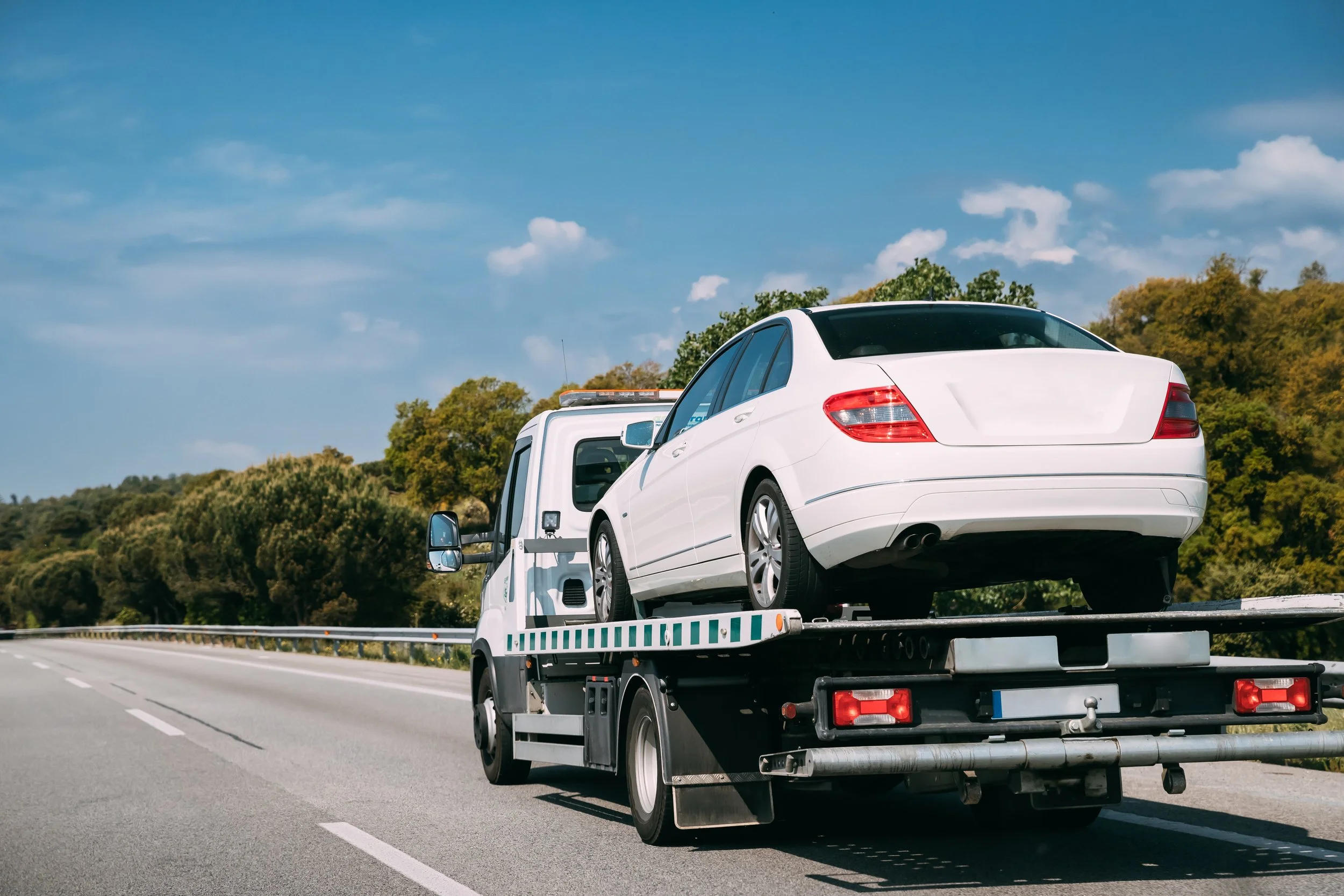 A white sedan car being transported on a flatbed tow truck on the highway with trees and a blue sky with clouds in the background.