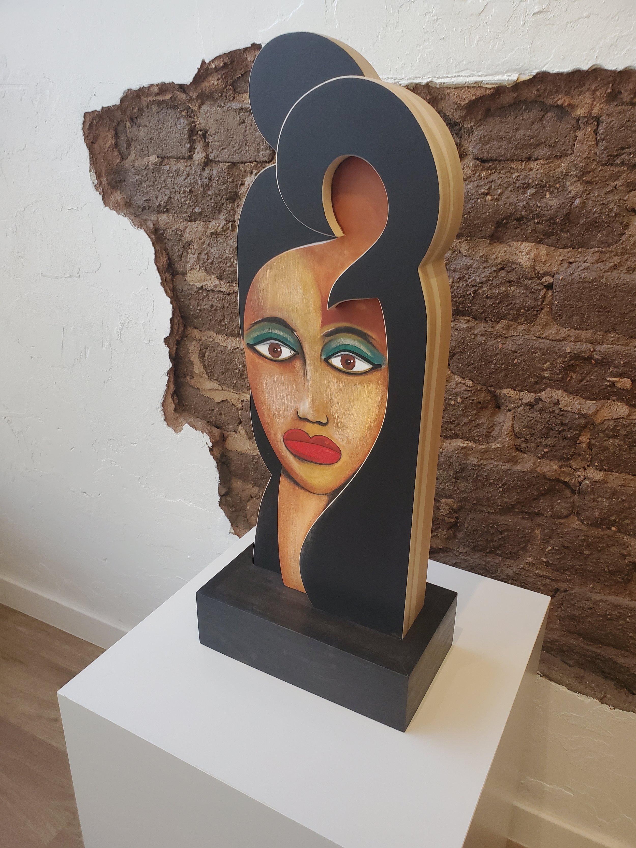 A tabletop wood sculpture featuring a painted face with exaggerated makeup and red lips, mounted on a black base. The sculpture is displayed against a brick wall.