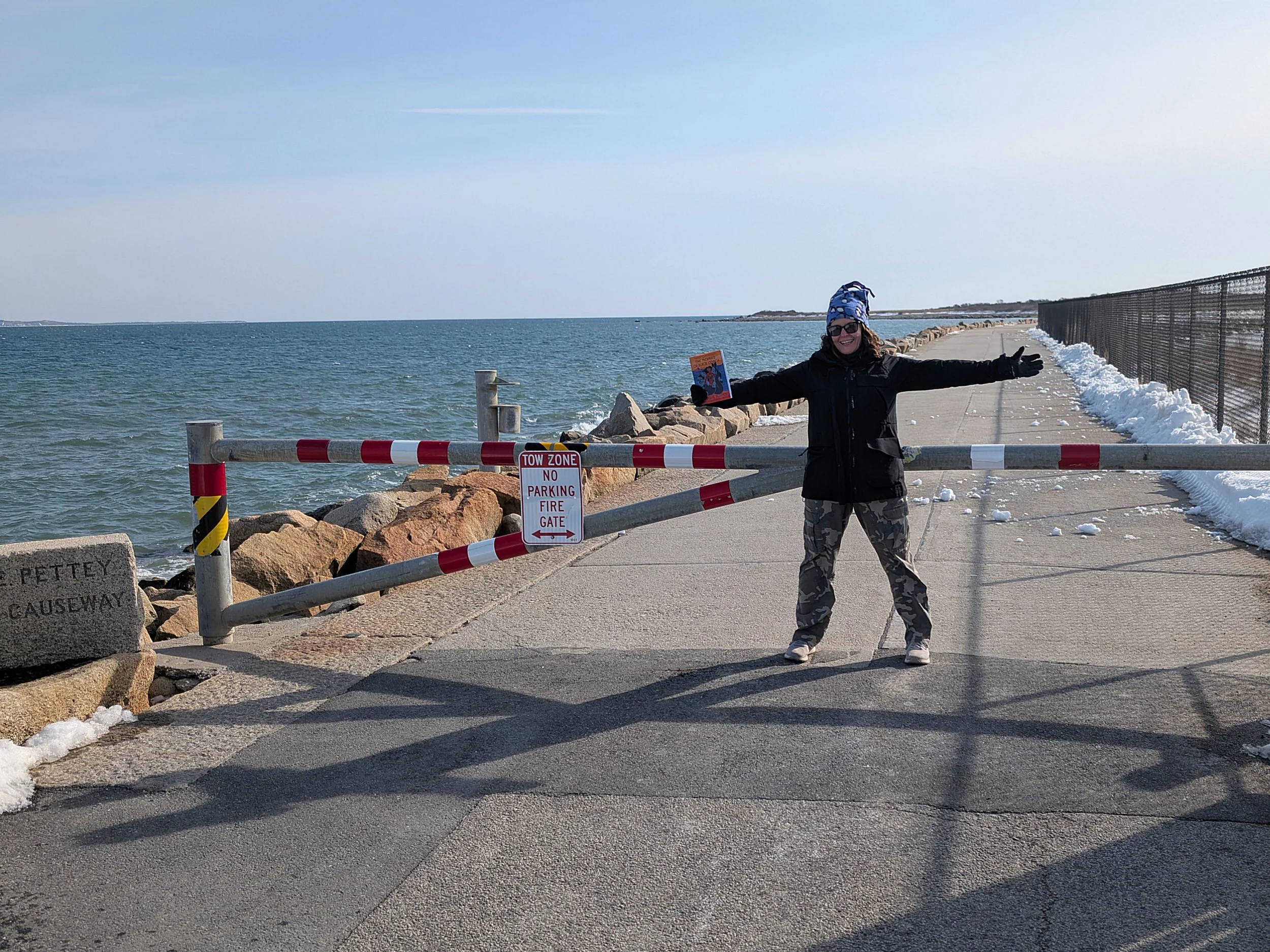 Krista Allen standing with arms open holding a book in front of the causeway gate.