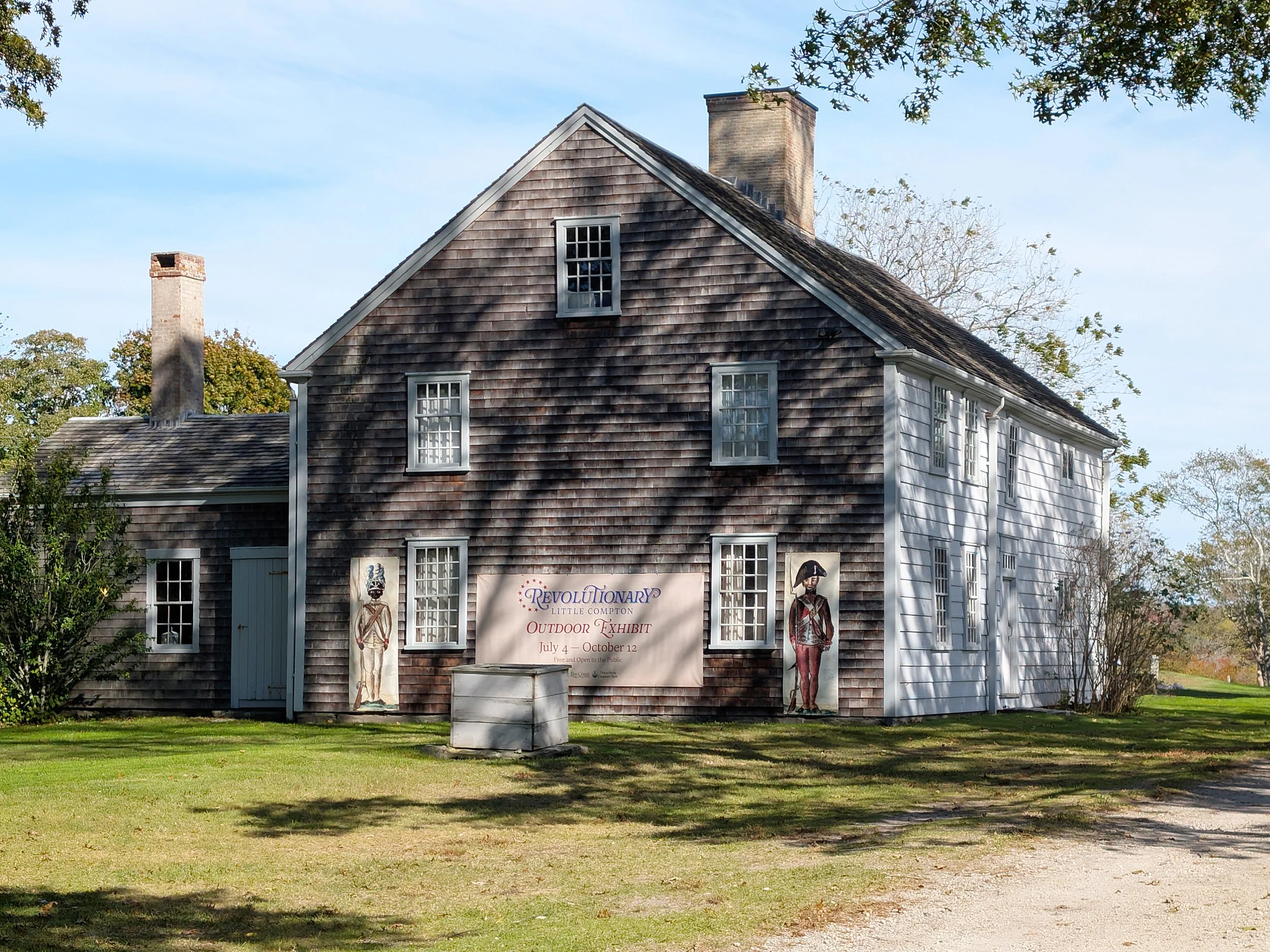 Little Compton Historical Society, a historic shingled house displaying an outdoor exhibit banner and life-size historical figures on the lawn. Image