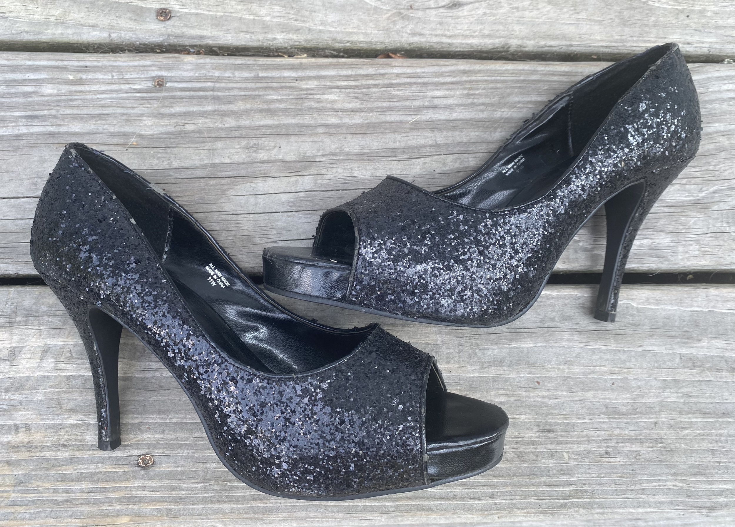 A pair of black glitter-covered peep-toe high-heeled shoes displayed on a wooden surface.