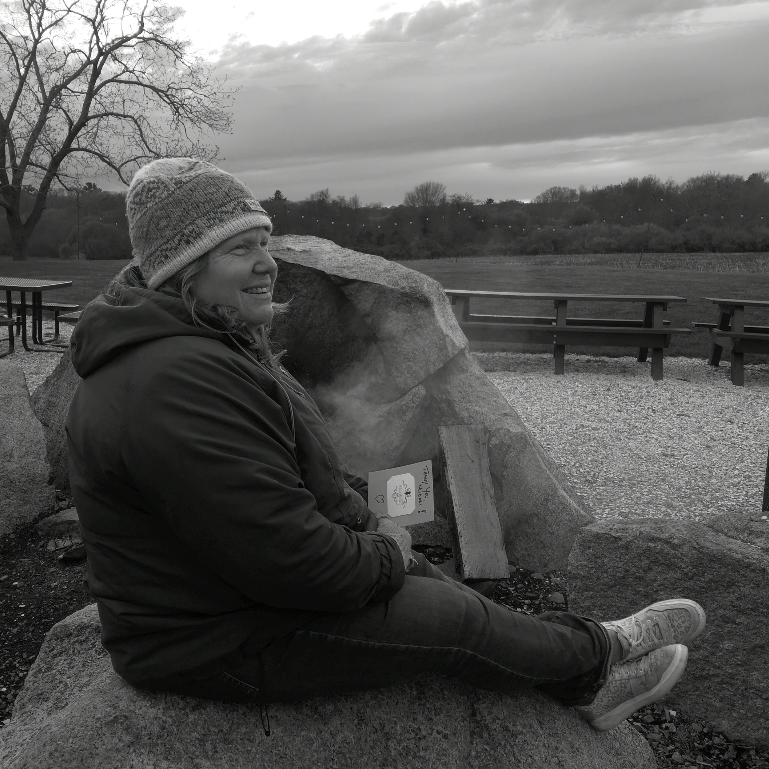 Black-and-white photograph of Midori Evans sitting outdoors on a bench, wearing a knit hat and coat, with an open landscape in the background.