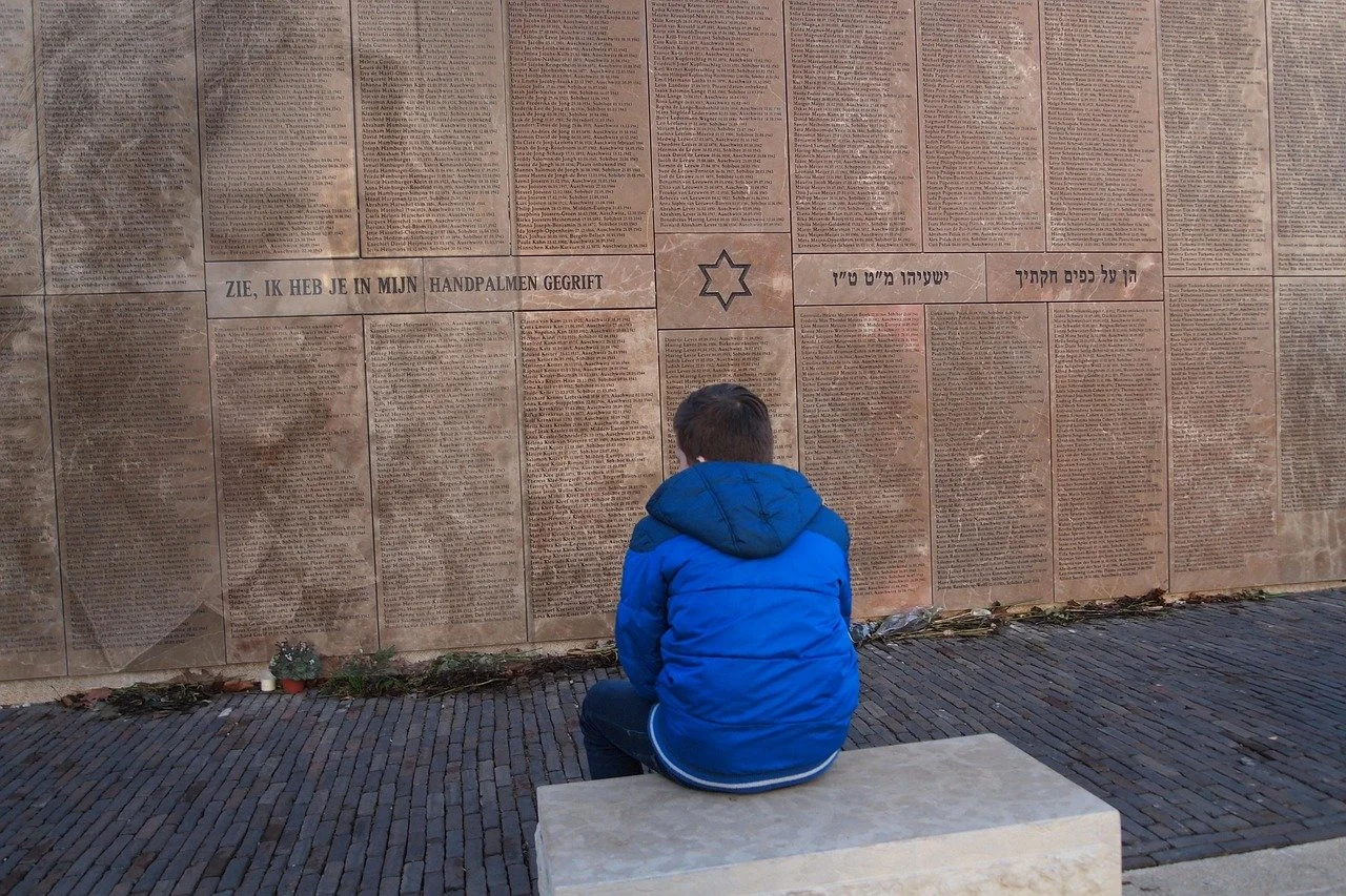 A person in a blue jacket sitting on a bench, facing a large memorial wall covered with engraved text.