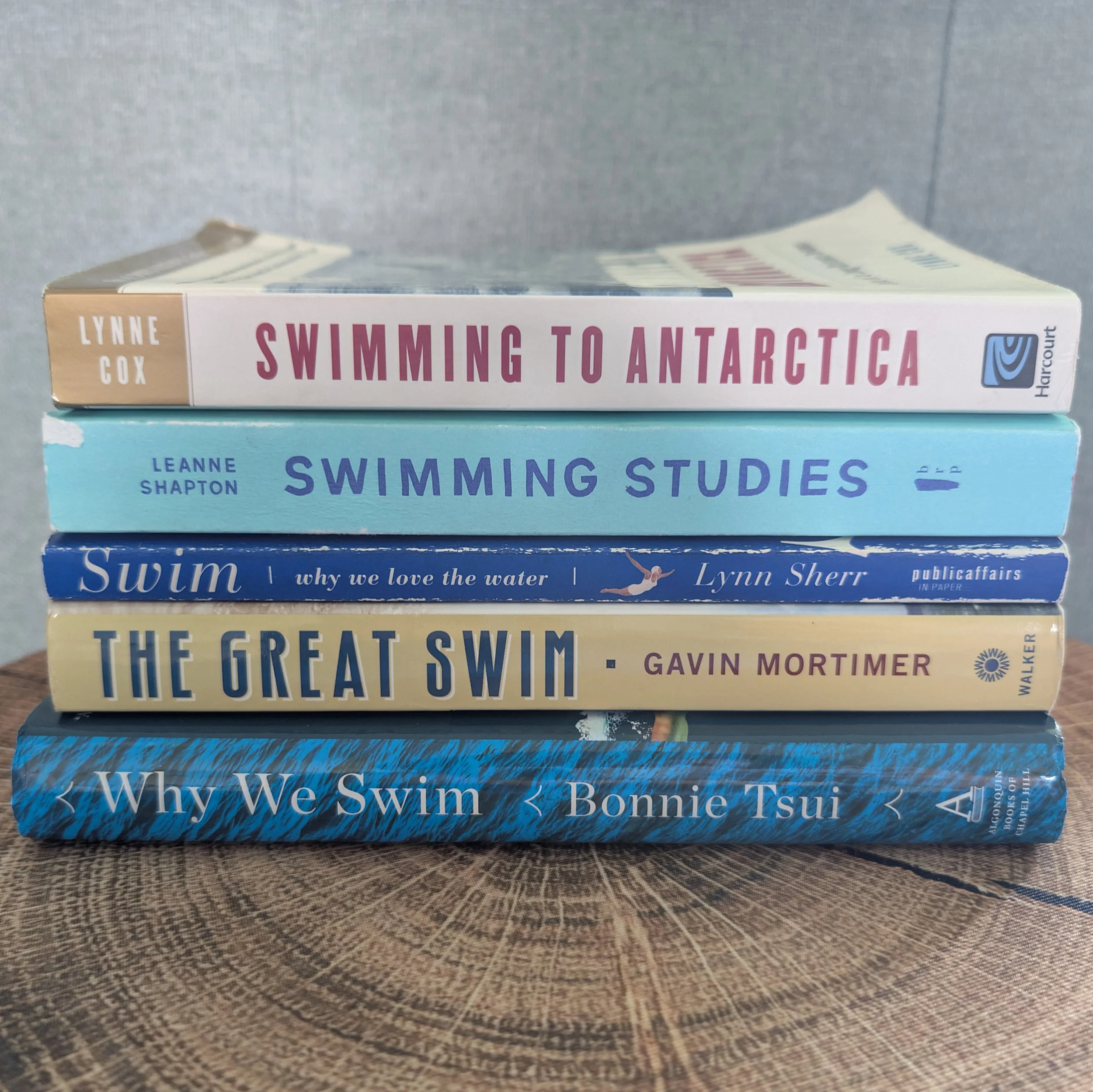 A stack of five books about swimming, including Swimming to Antarctica, Swimming Studies, Swim, The Great Swim, and Why We Swim, arranged on a wooden surface.