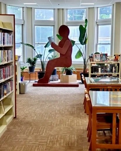 A library interior with bookshelves and reading tables featuring a large reddish silhouette sculpture of a seated figure in reading position
