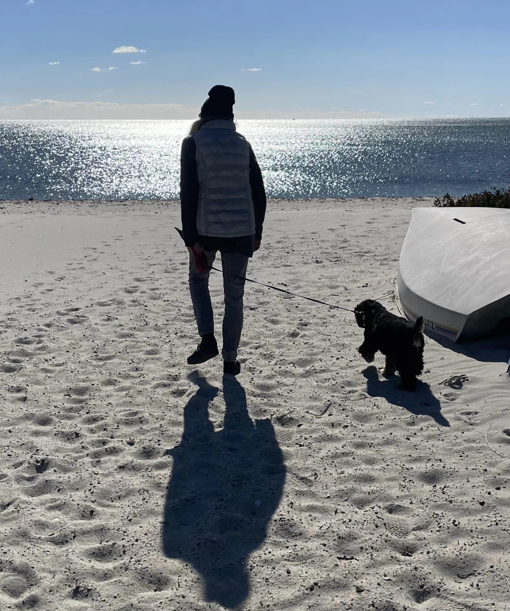 A woman walks on a sandy beach with a small black dog on a leash