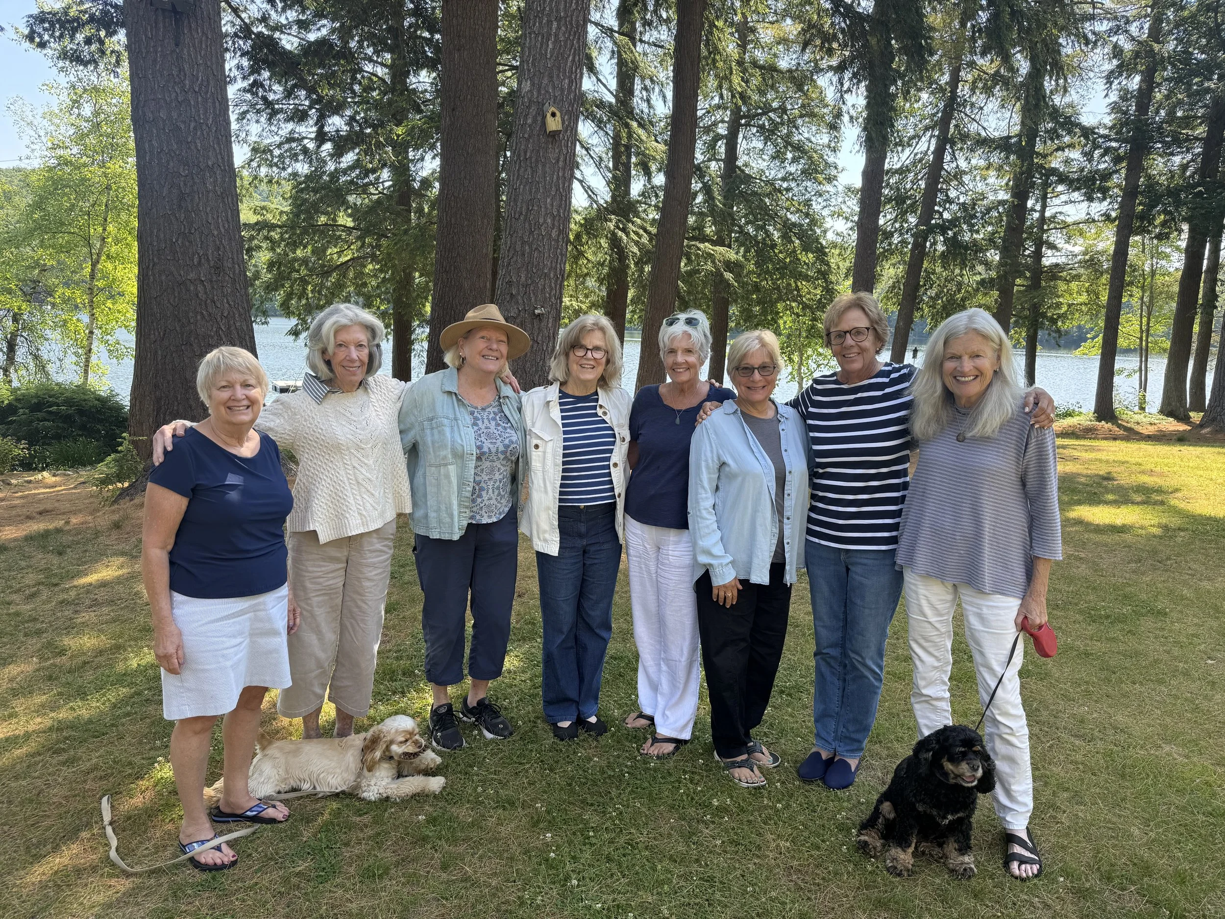 Eight women pose together outdoors near a lake, with two small spaniel dogs sitting on the grass in front of them.