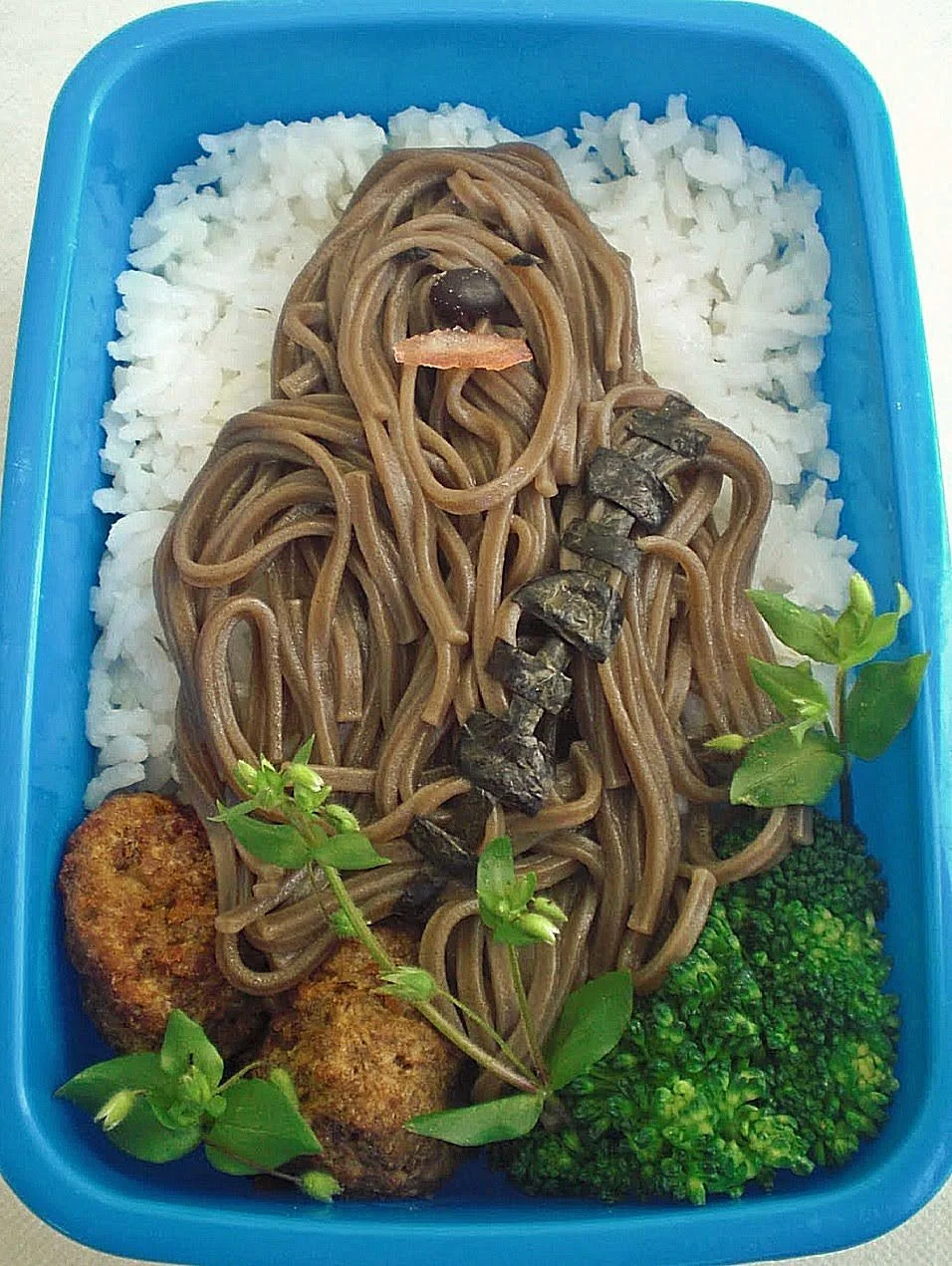 A blue lunch container filled with white rice topped with brown noodles arranged to look like a long-haired person.