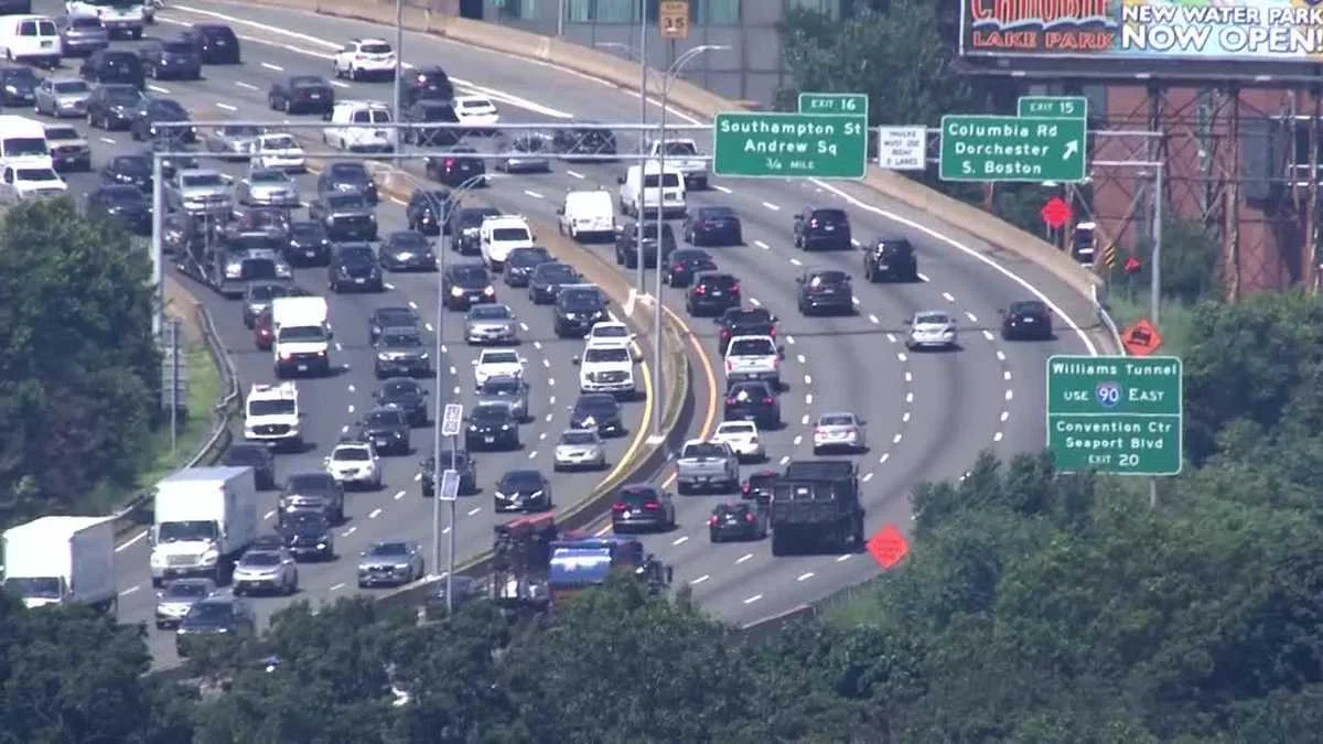 Heavy traffic on a multi-lane highway with cars and trucks approaching an exit, visible road signs, and surrounding greenery.