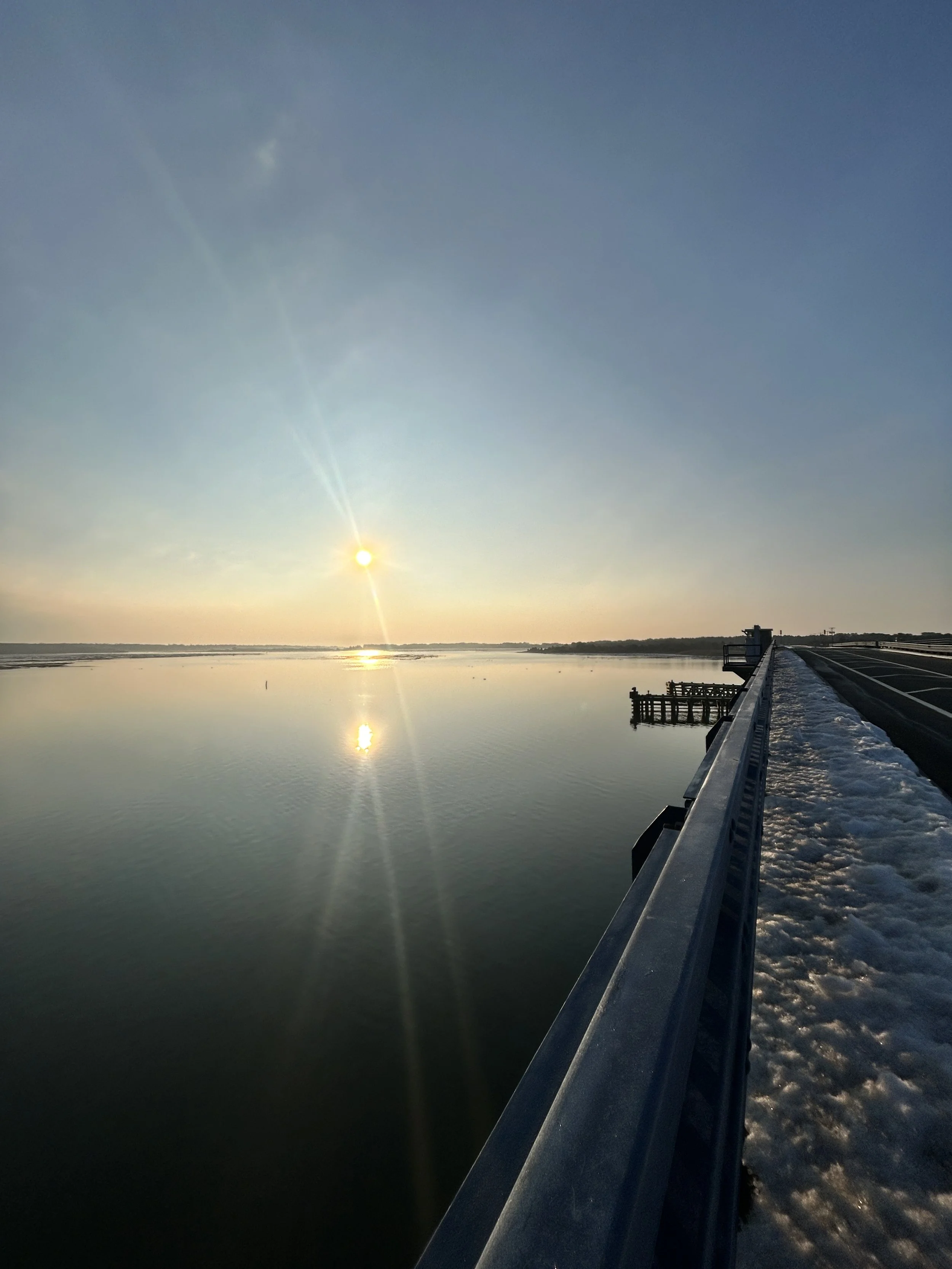 A picture of a sunset over a calm body of water. A long railing extends from the bottom right toward the center, with a dusting of snow visible on its edge.