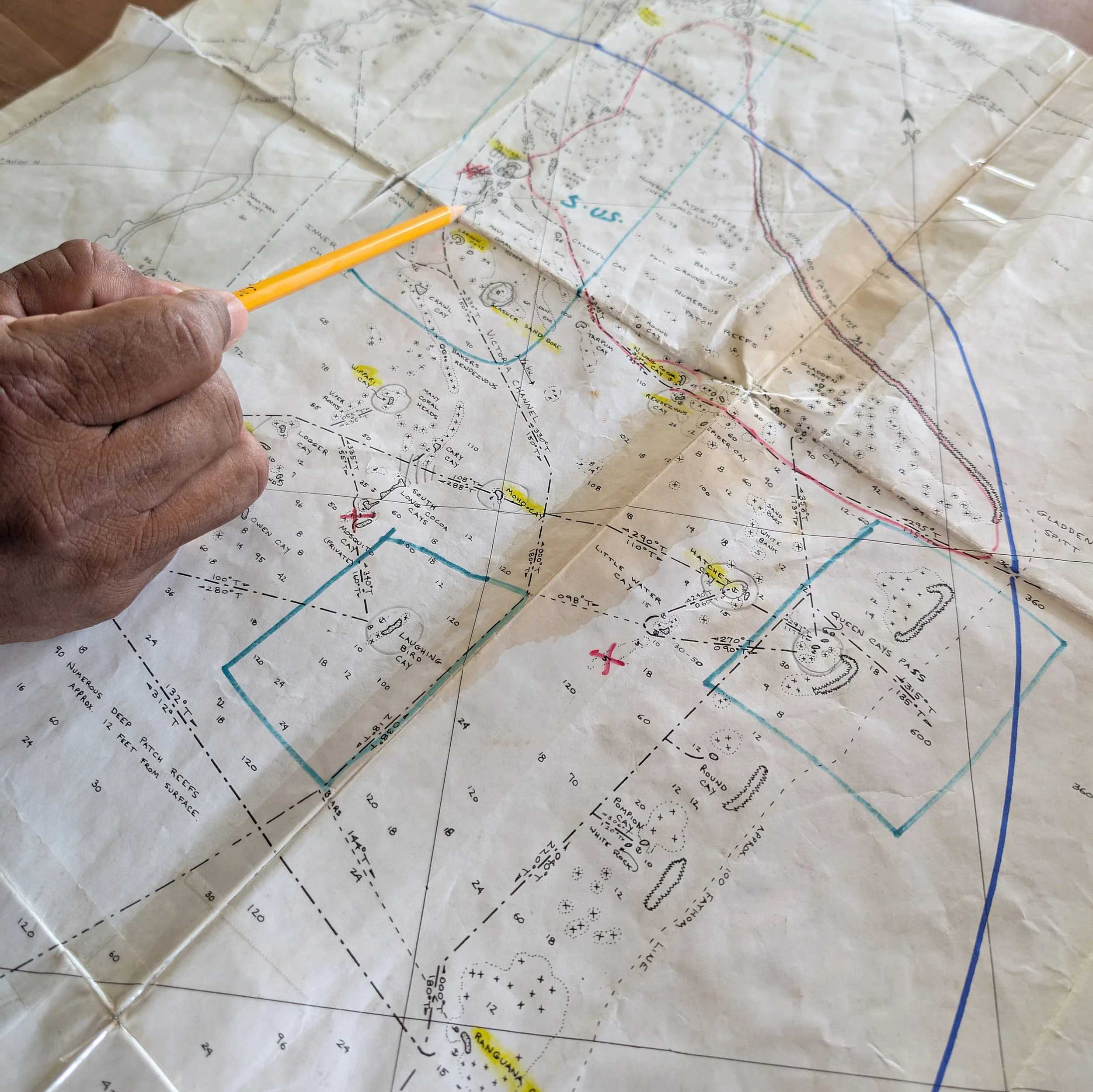 Close-up of a nautical chart with a person pointing to a location using a yellow pencil.
