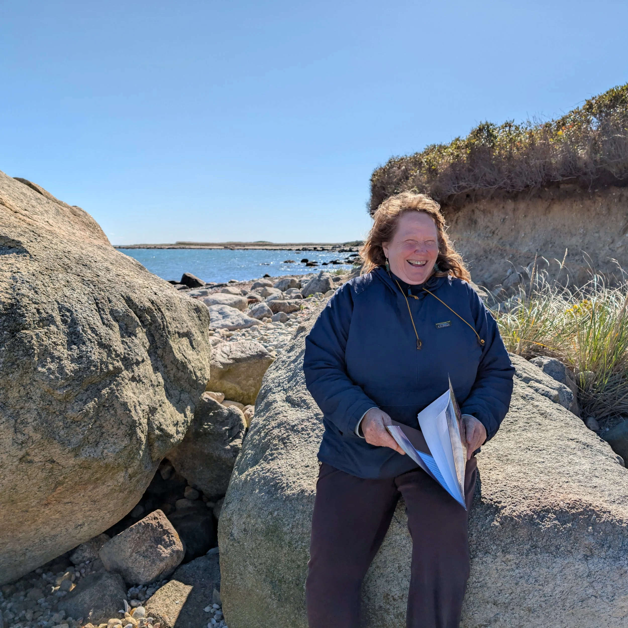 Midori Evans standing on coastal rocks holding papers with the ocean and shoreline in the background