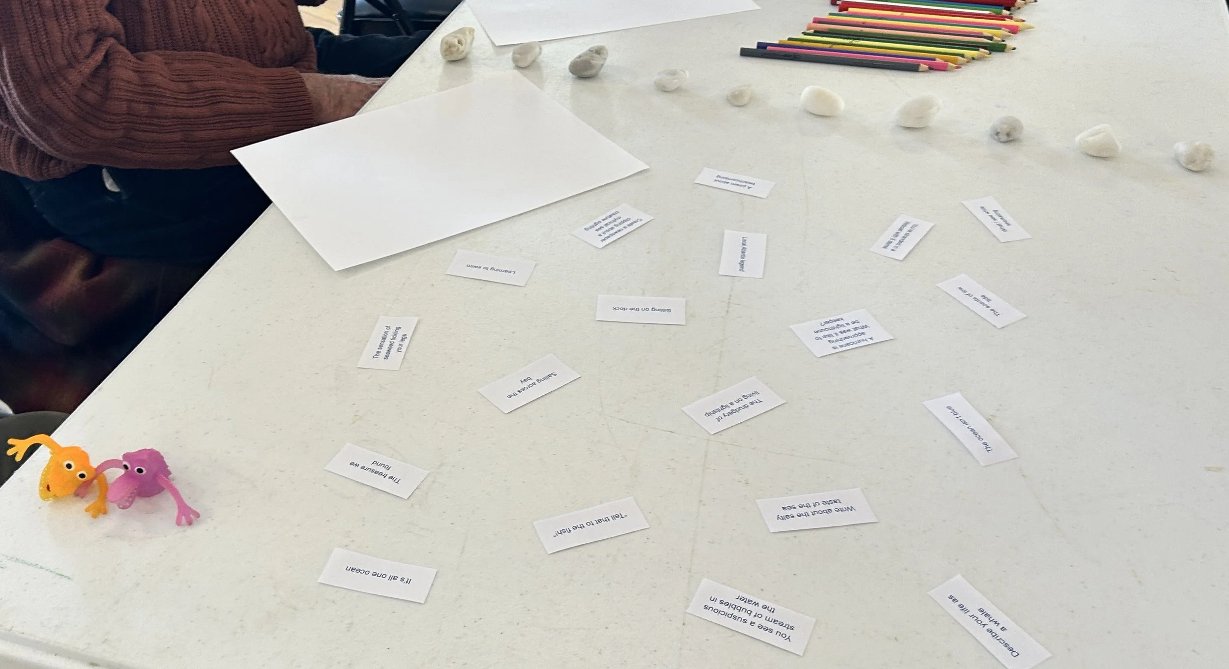small white slips of paper with text scattered around, colorful toy figurines, a row of small stones, a stack of colored pencils, and a blank sheet of paper on a white table used for an interactive activity