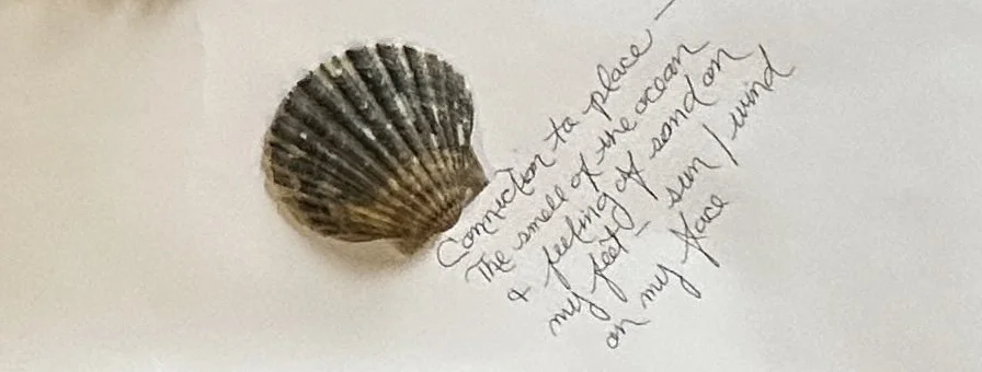 A close-up of a scalloped seashell resting on a white surface next to handwritten text in cursive. The text reads: "Connection to place—The smell of the ocean + feeling of sand on my feet—sun / wind on my face."