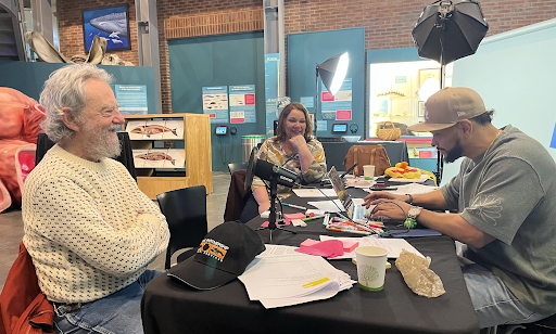 Three people sit around a table in a studio setting with microphones and papers discussing over something during a casual work session