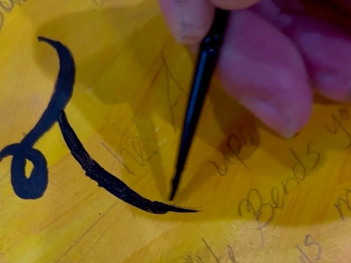A hand creates a calligraphy stroke with a black pen on yellow paper.