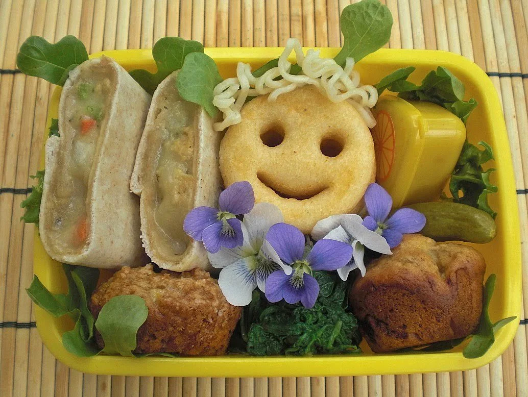A yellow bento box arranged with a smiley-face potato, wraps, greens, small muffins, and edible flowers