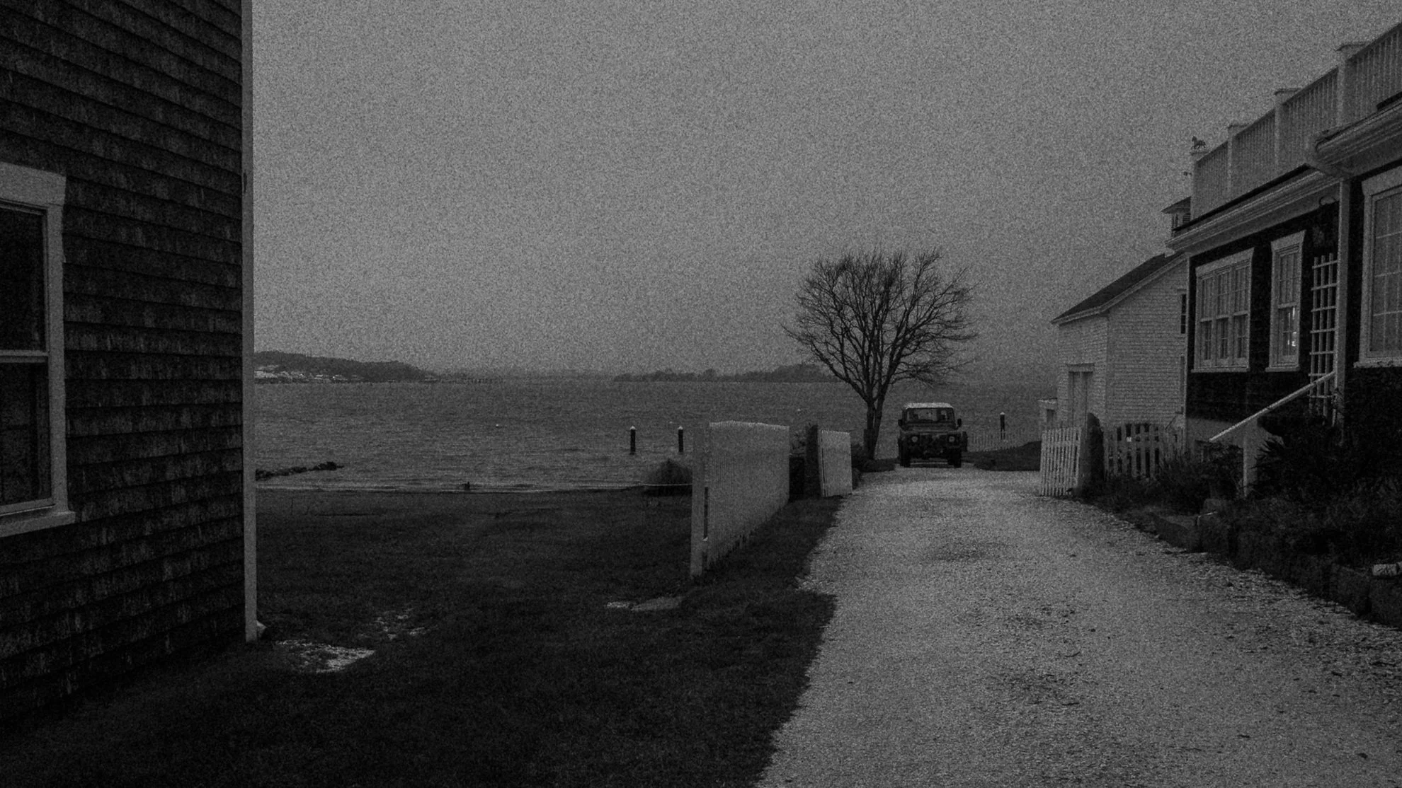 A black-and-white view of a gravel road leading toward the waterfront, with houses on either side.