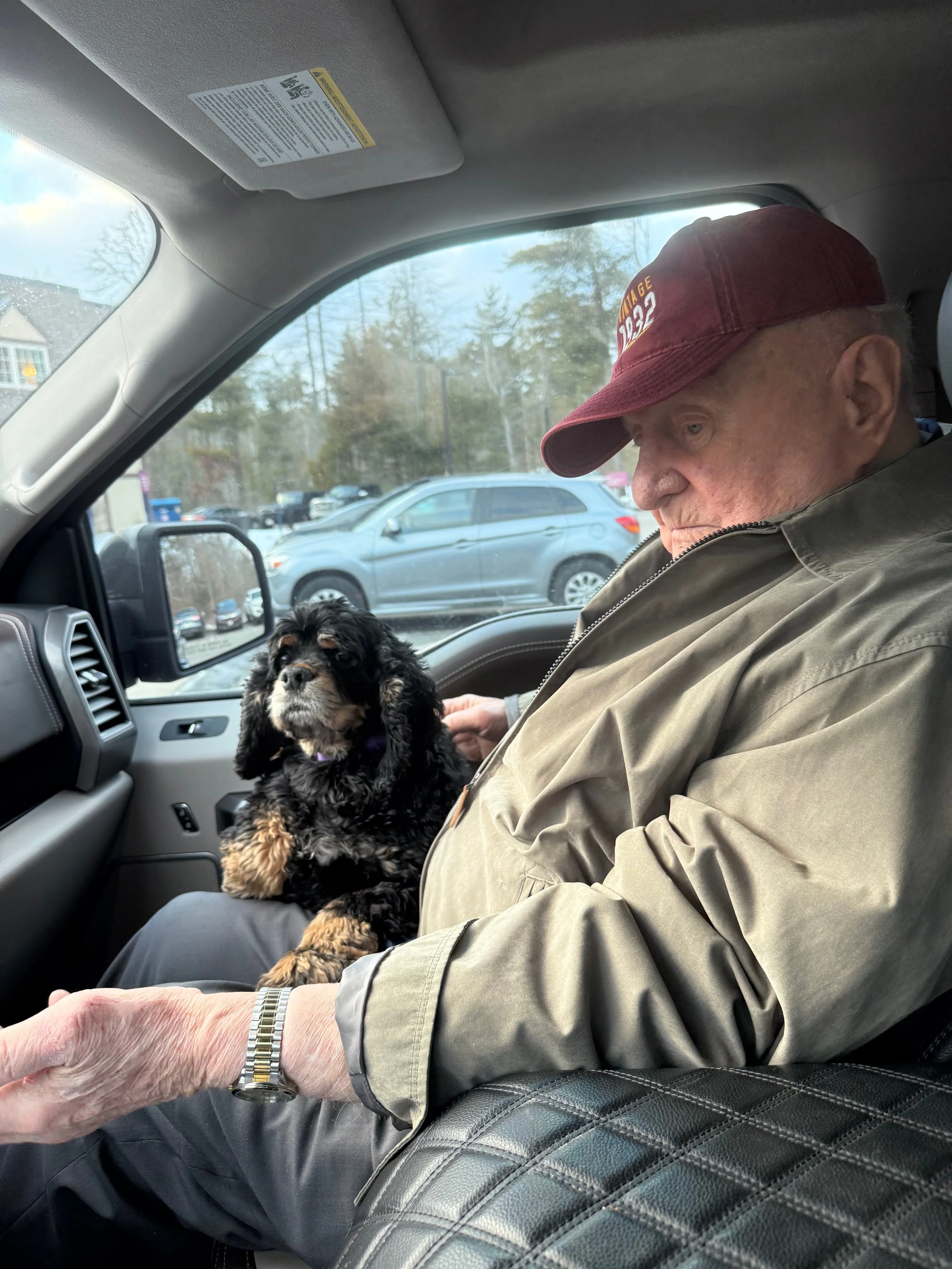 An older man sits in the driver’s seat of a car beside a black-and-tan spaniel sitting on his lap
