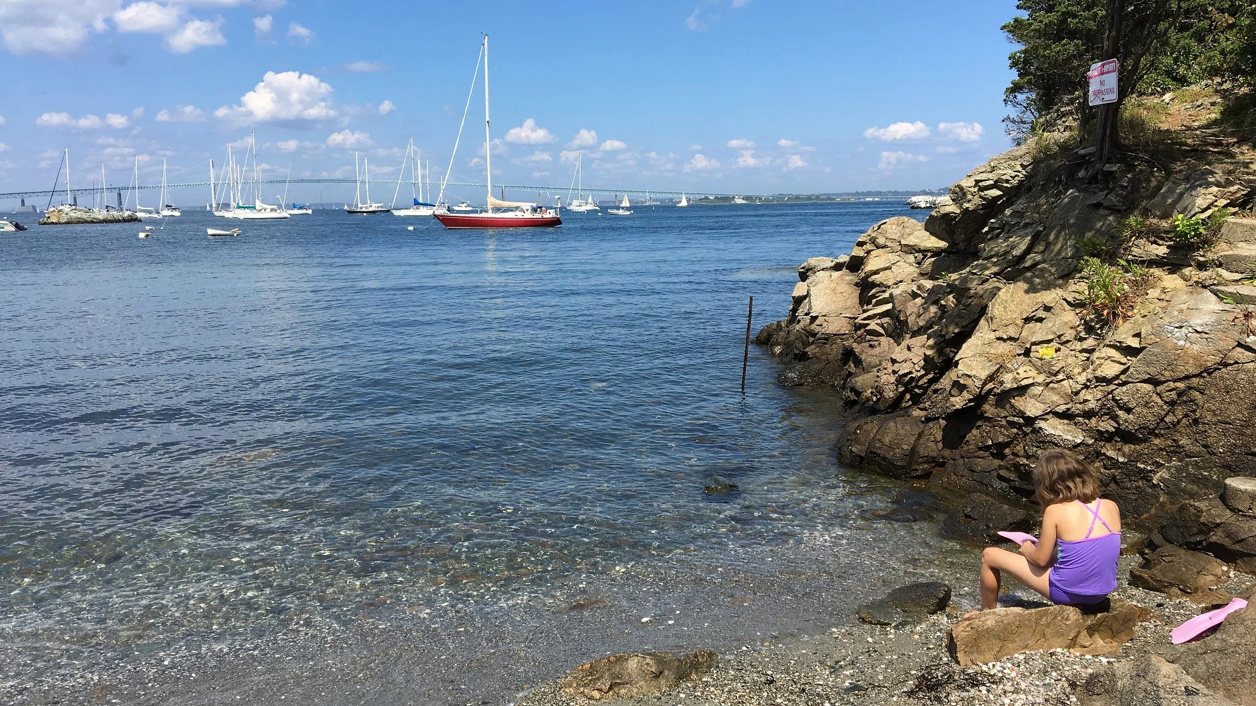 A girl in a purple swimswear sits on rocks beside a calm coastal inlet, with sailboats in the water and rocky cliffs along the shore.
