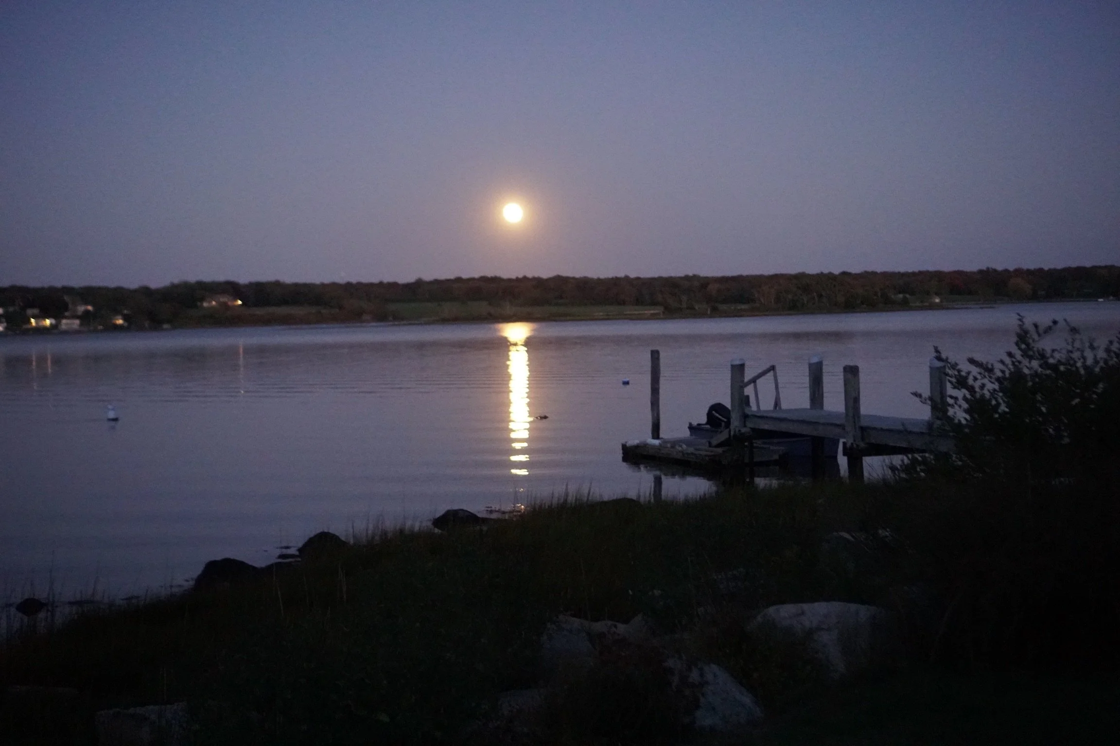 Full moon reflecting on calm water beside a small dock at dusk.