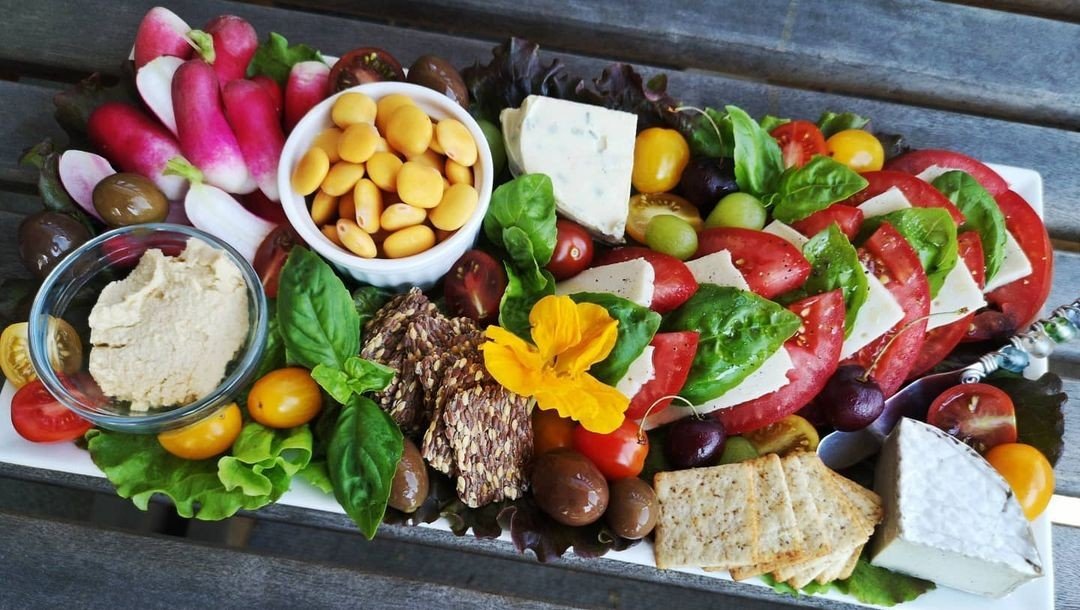 A large platter filled with colorful fresh foods including sliced tomatoes, basil leaves, cheeses, olives, crackers, radishes, beans, and dips