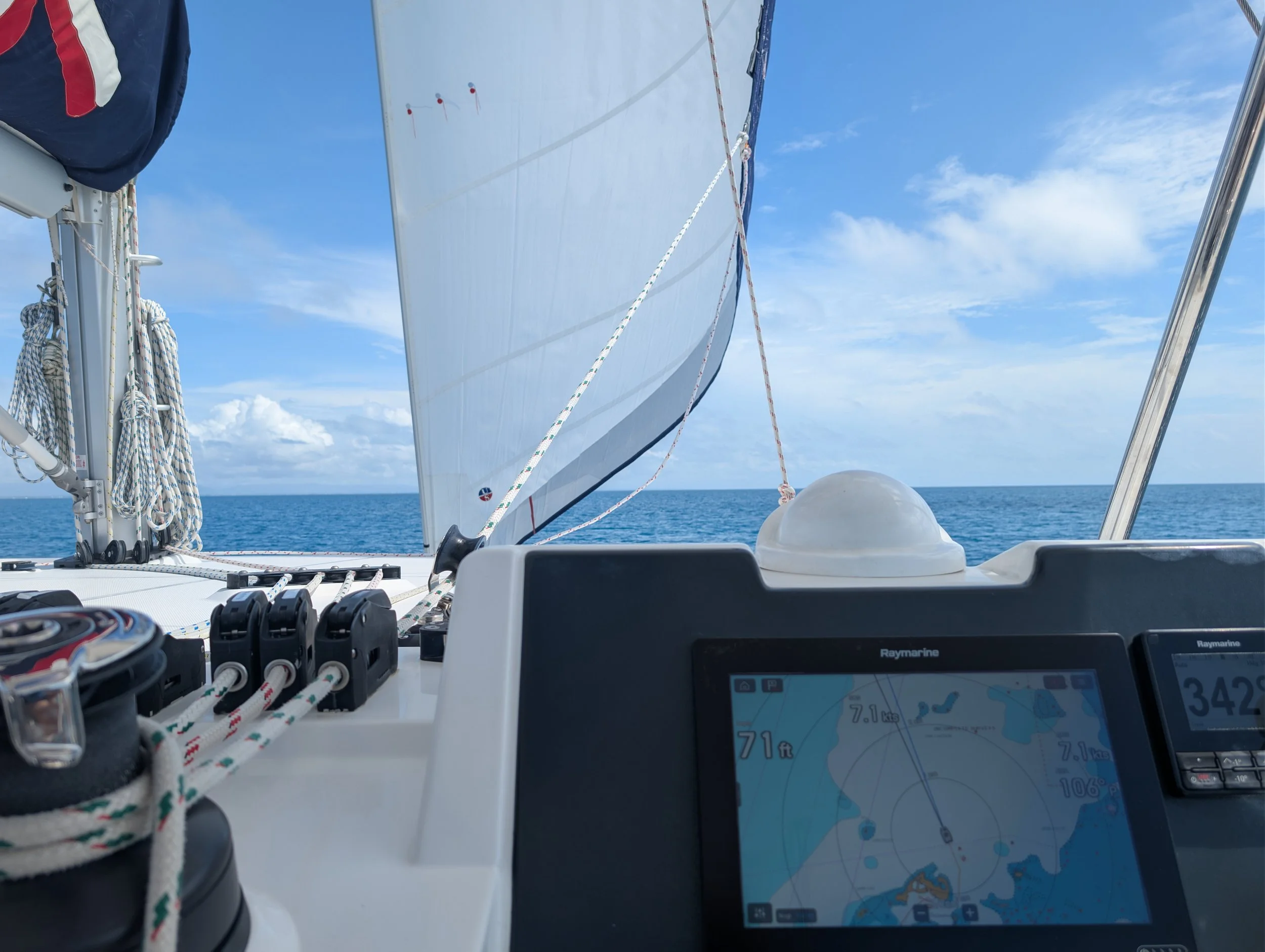 View from a sailboat with a raised white sail, navigation displays, and open blue ocean