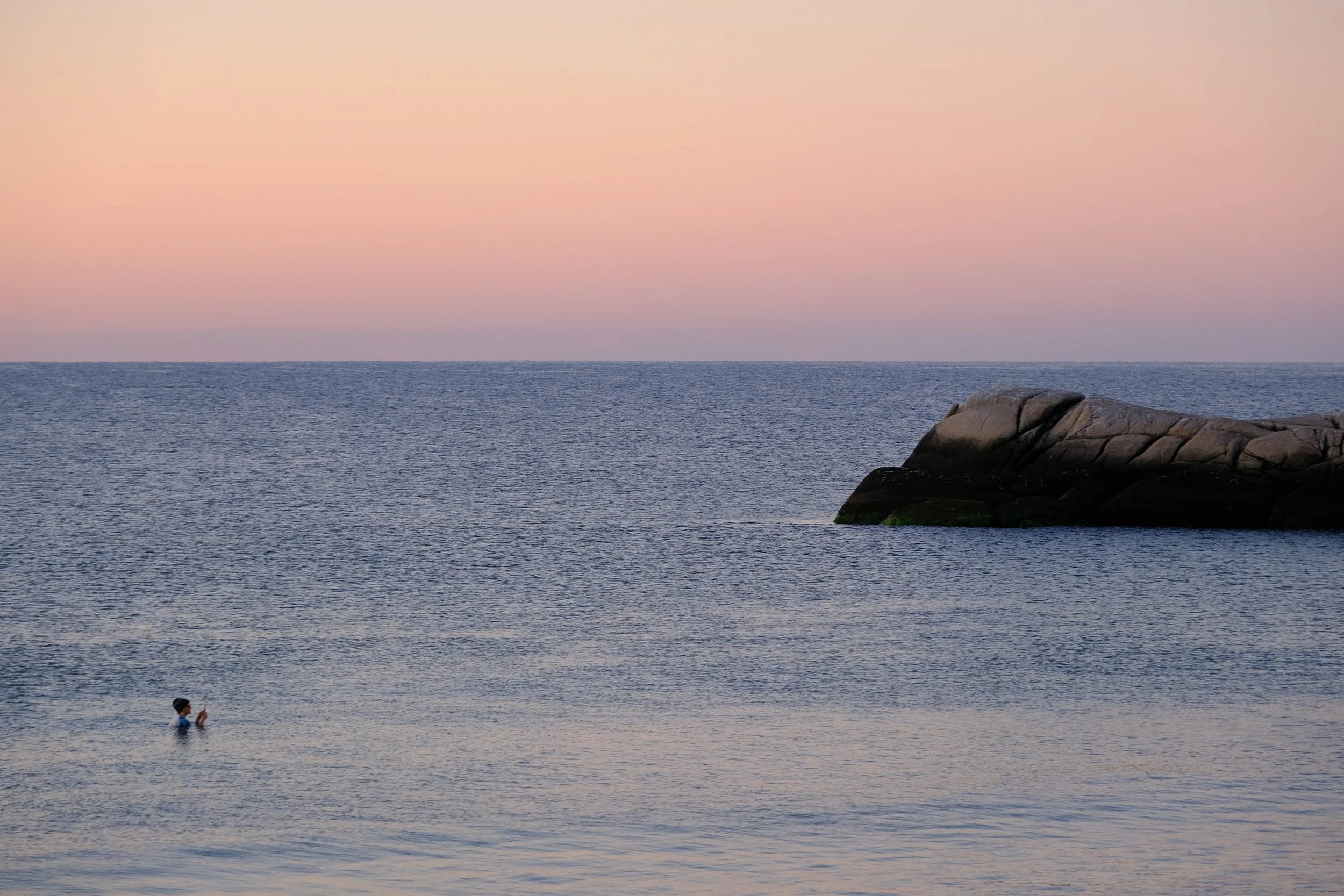 A calm ocean at sunset with soft pink skies, a rocky outcrop, and a lone swimmer floating near the shore.