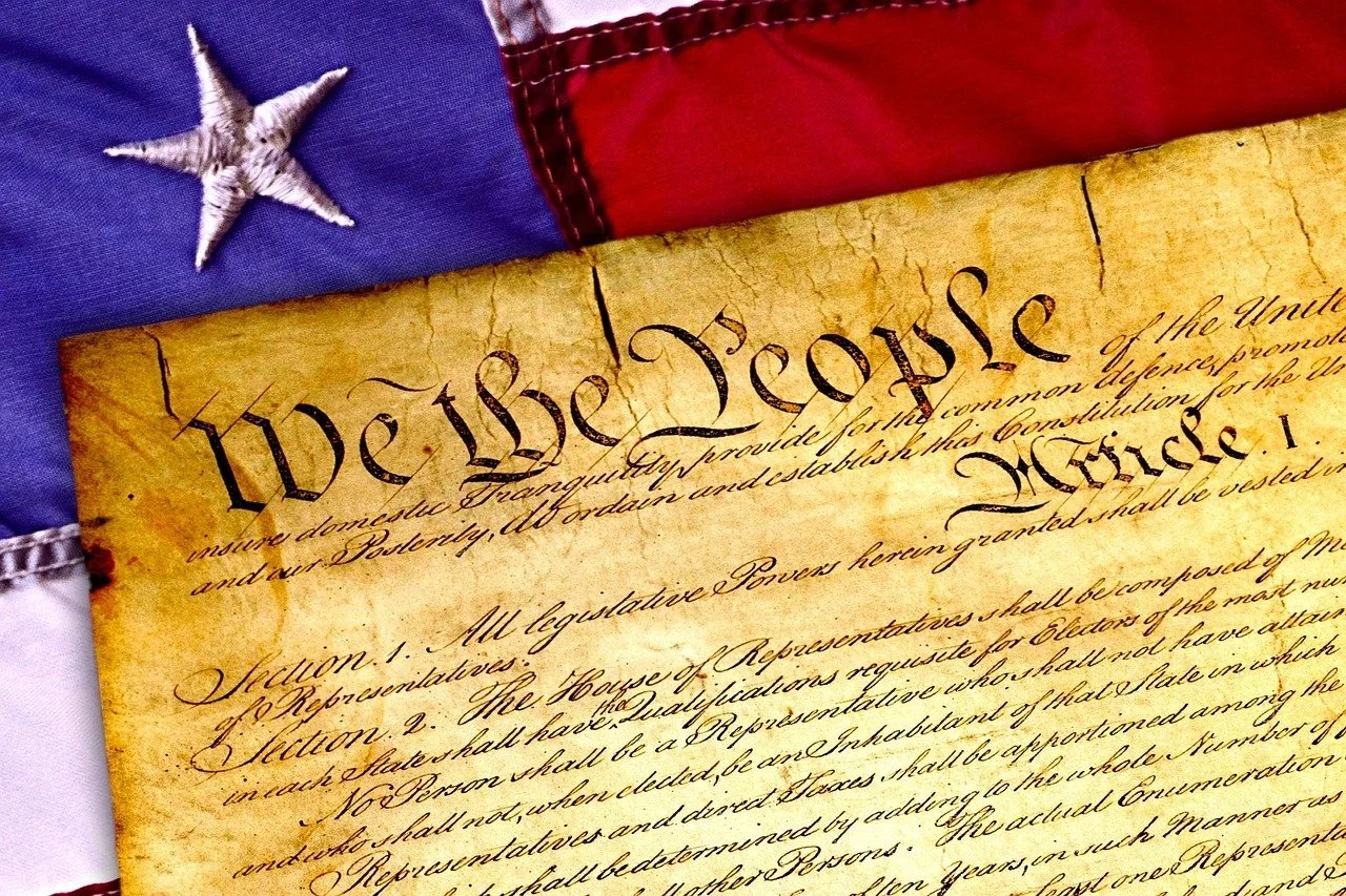 A close-up of the opening text “We the People” from the U.S. Constitution on aged parchment, partially overlaid with the American flag.