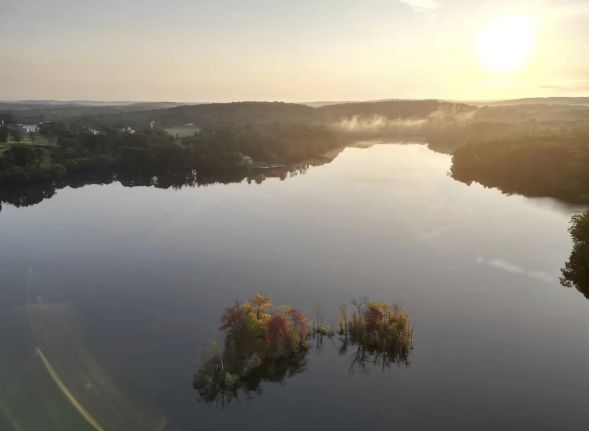A view of a calm lake at sunrise, with soft mist rising above the water.