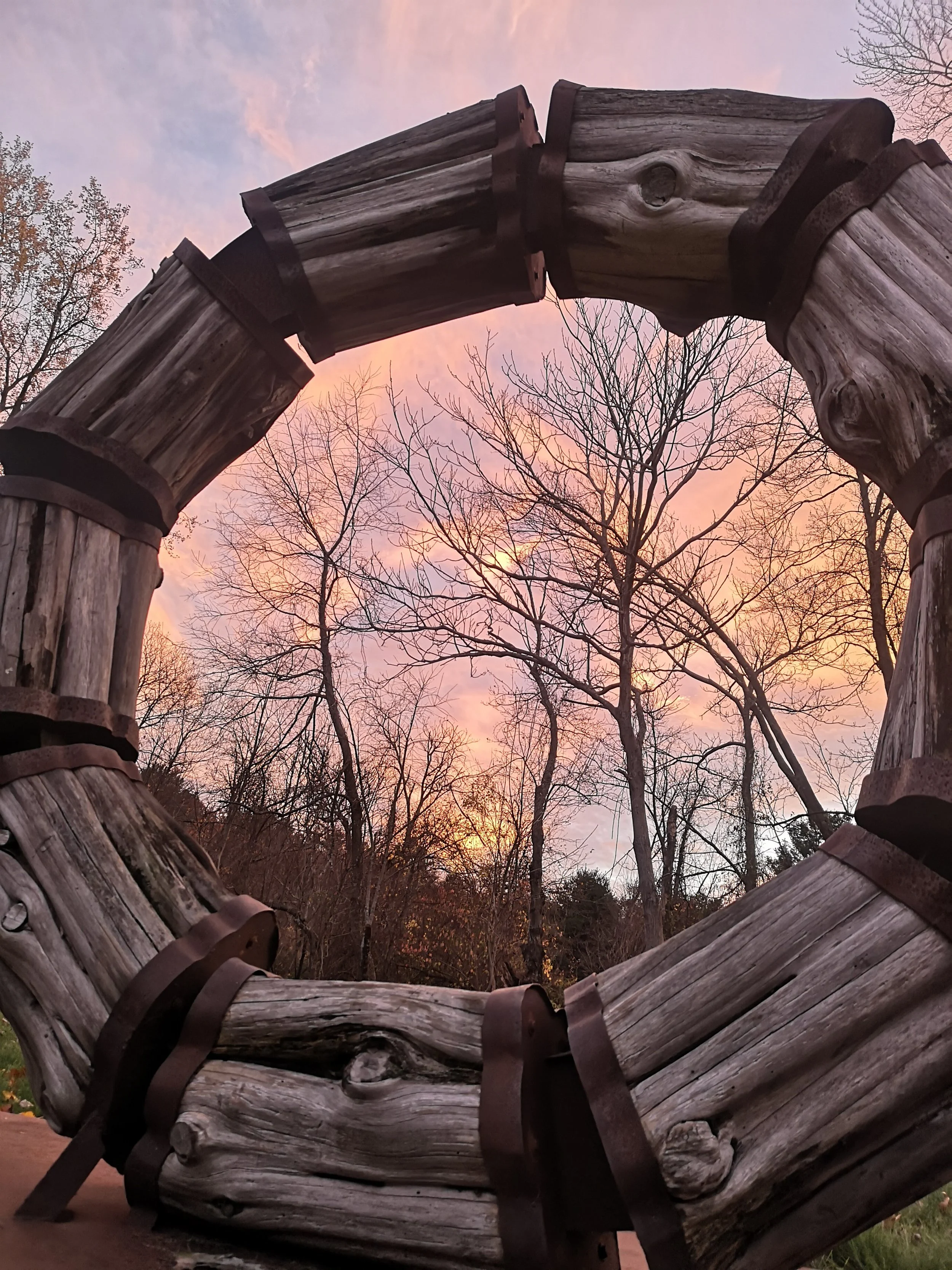 pink sunset and trees visible through a round sculpture of large wooden logs