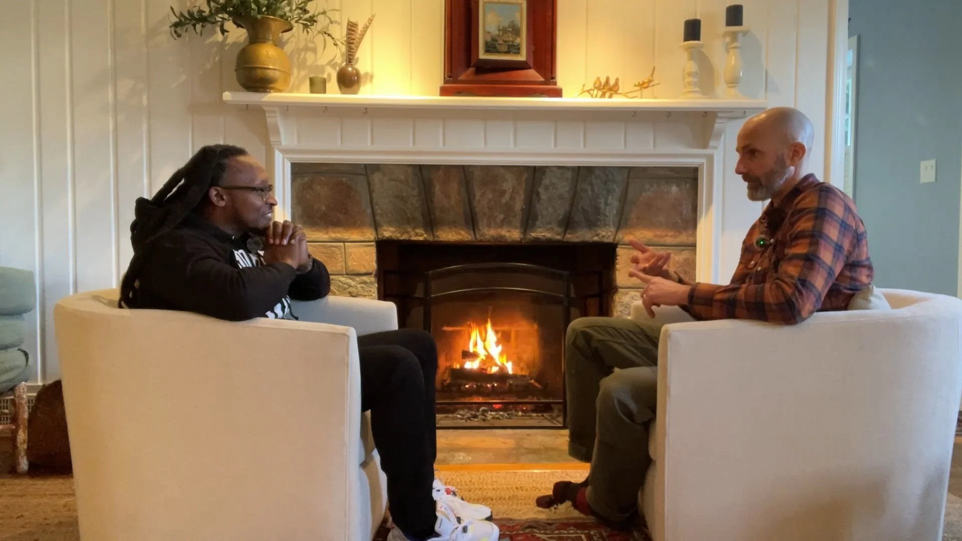 Two men sitting in white armchairs around a lit fireplace, engaged in conversation inside a cozy living room with decorative items on the mantel.