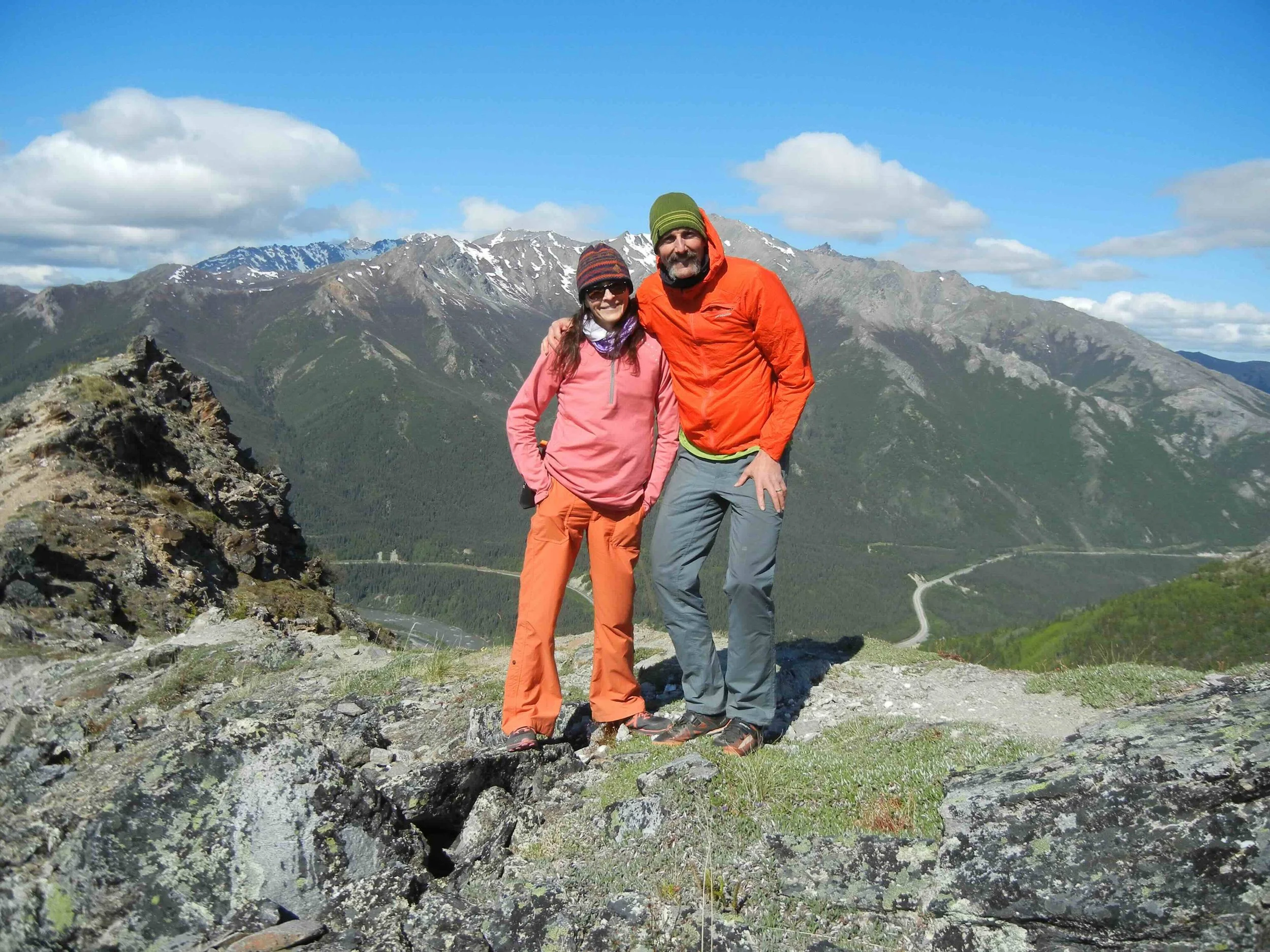 Two hikers, a woman in a pink jacket and orange pants and a man in an orange jacket and gray pants, standing on a rocky ledge with mountains and a partly cloudy sky in the background.